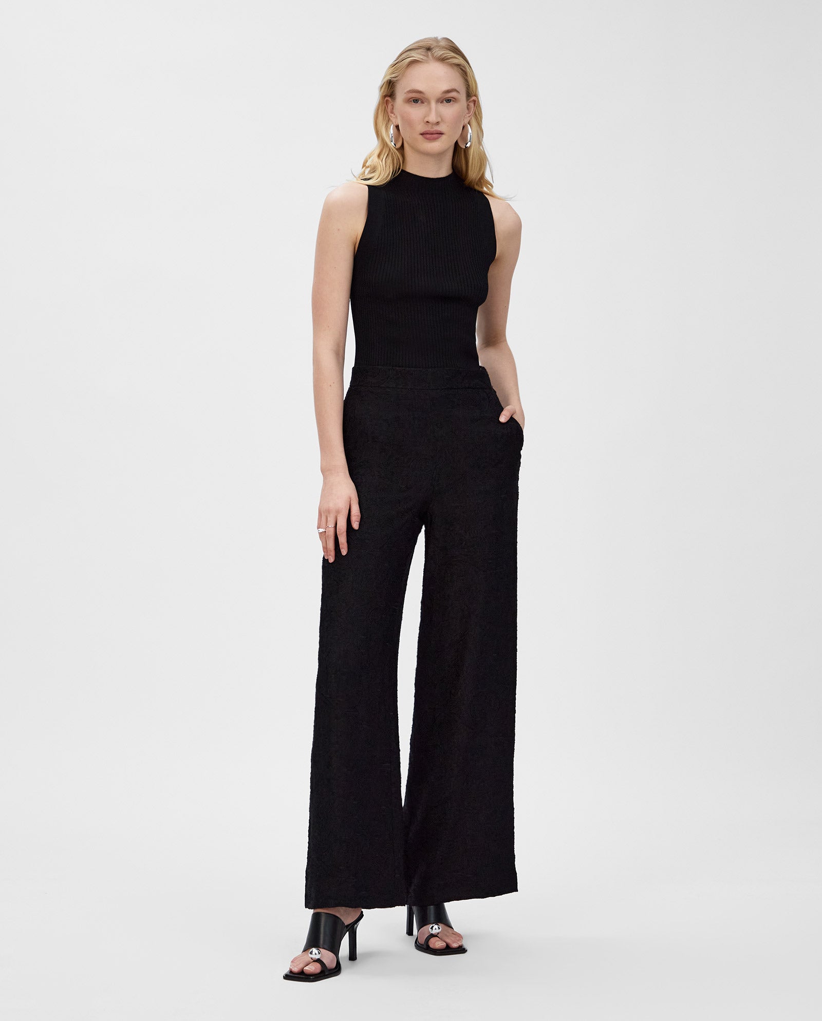 A woman wears the IVY OAK KESSIE sleeveless black top and wide-leg black pants with black heeled sandals, standing on a white background.