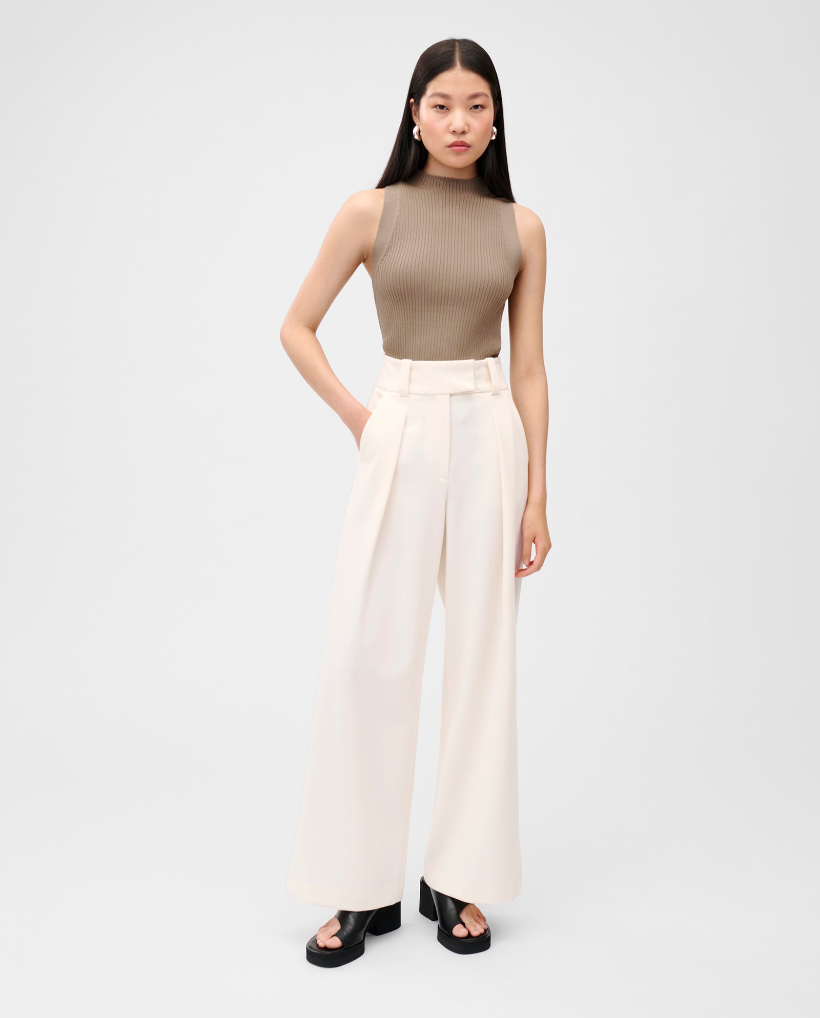 A woman wears the IVY OAK KESSIE Top, a sleeveless brown piece in stretchy fabric, paired with high-waisted white wide-leg pants and black platform sandals.