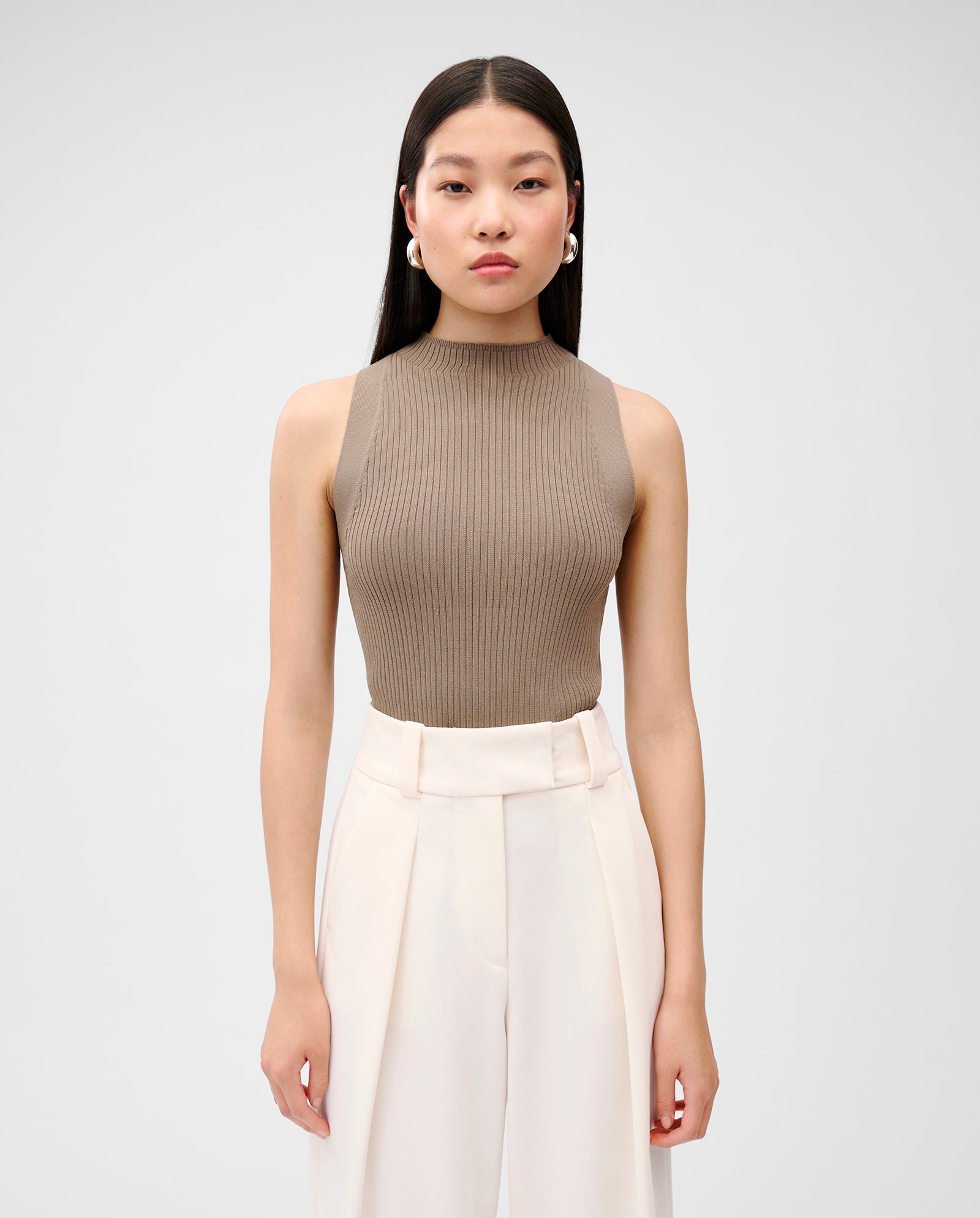 A woman with straight dark hair wears the KESSIE Top by IVY OAK, a precise fit sleeveless taupe top, paired with high-waisted white pants and stands facing forward.