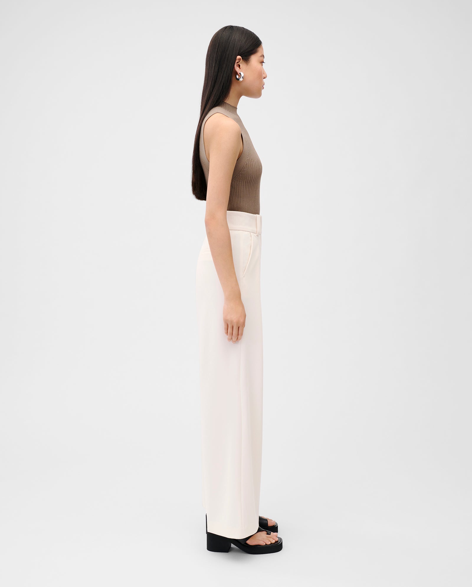 A woman stands in profile against a plain background, wearing the IVY OAK KESSIE brown strapless knit top, white wide-leg pants, and black sandals.