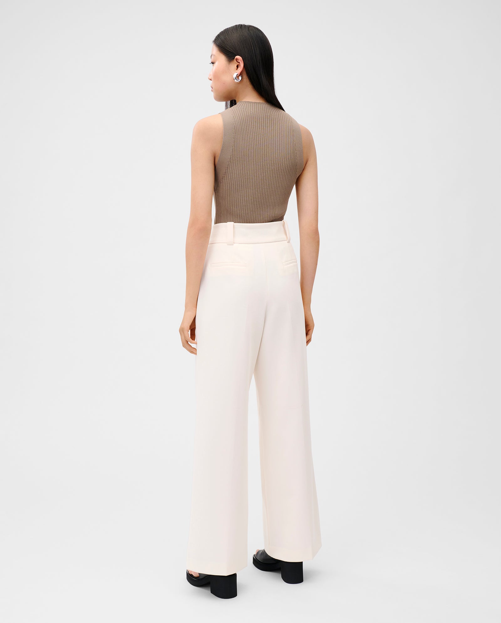A woman faces away wearing the IVY OAK KESSIE Top, a brown strapless knit piece with a precise fit, styled with wide-leg white pants and black platform sandals.