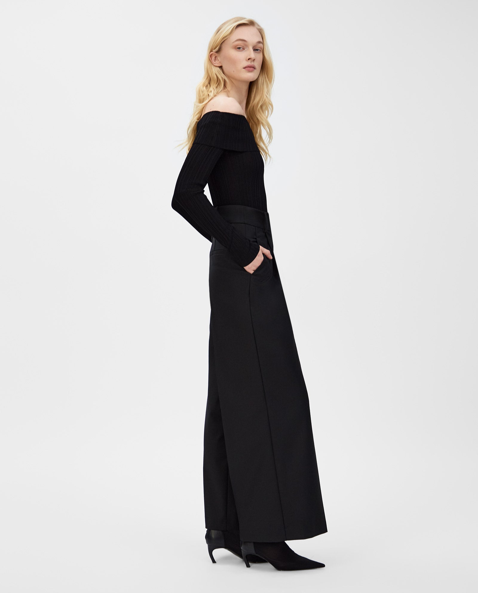 Woman wearing the IVY OAK KALLA Top in black with a carmen neckline and wide-leg pants, standing sideways with hands in pockets against a white background.