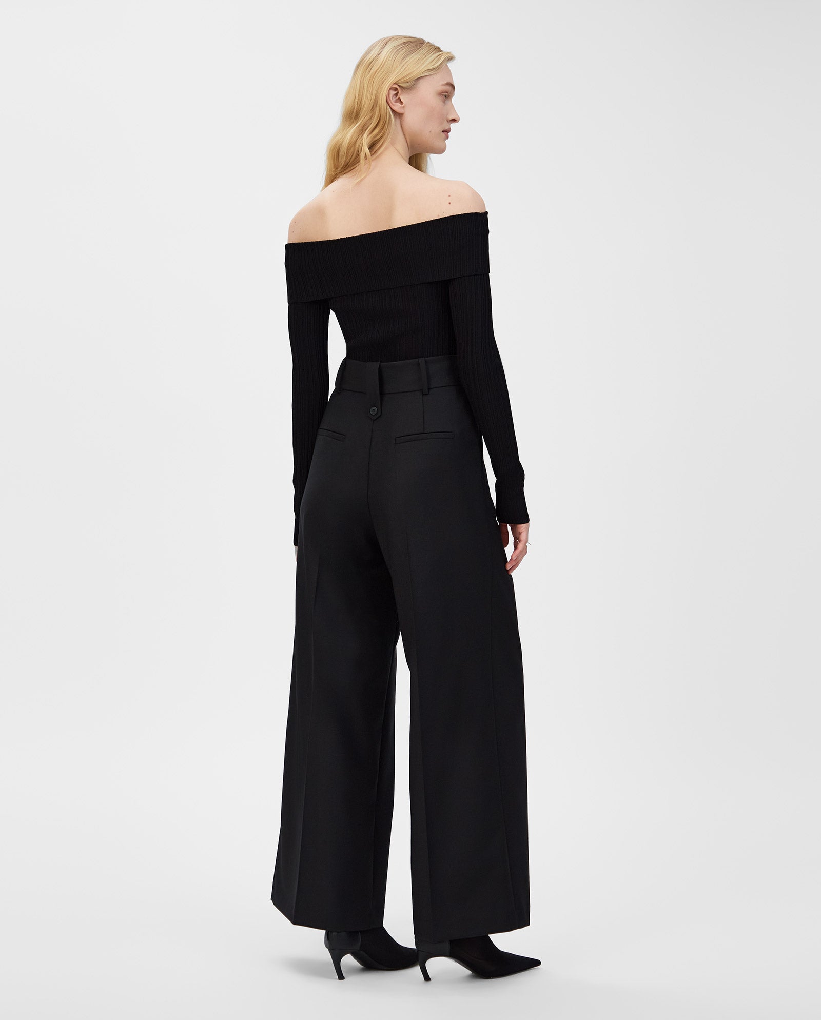 A woman stands against a plain background, wearing the IVY OAK KALLA Top in black with a carmen neckline and pairing it with wide-leg trousers, looking to the side.