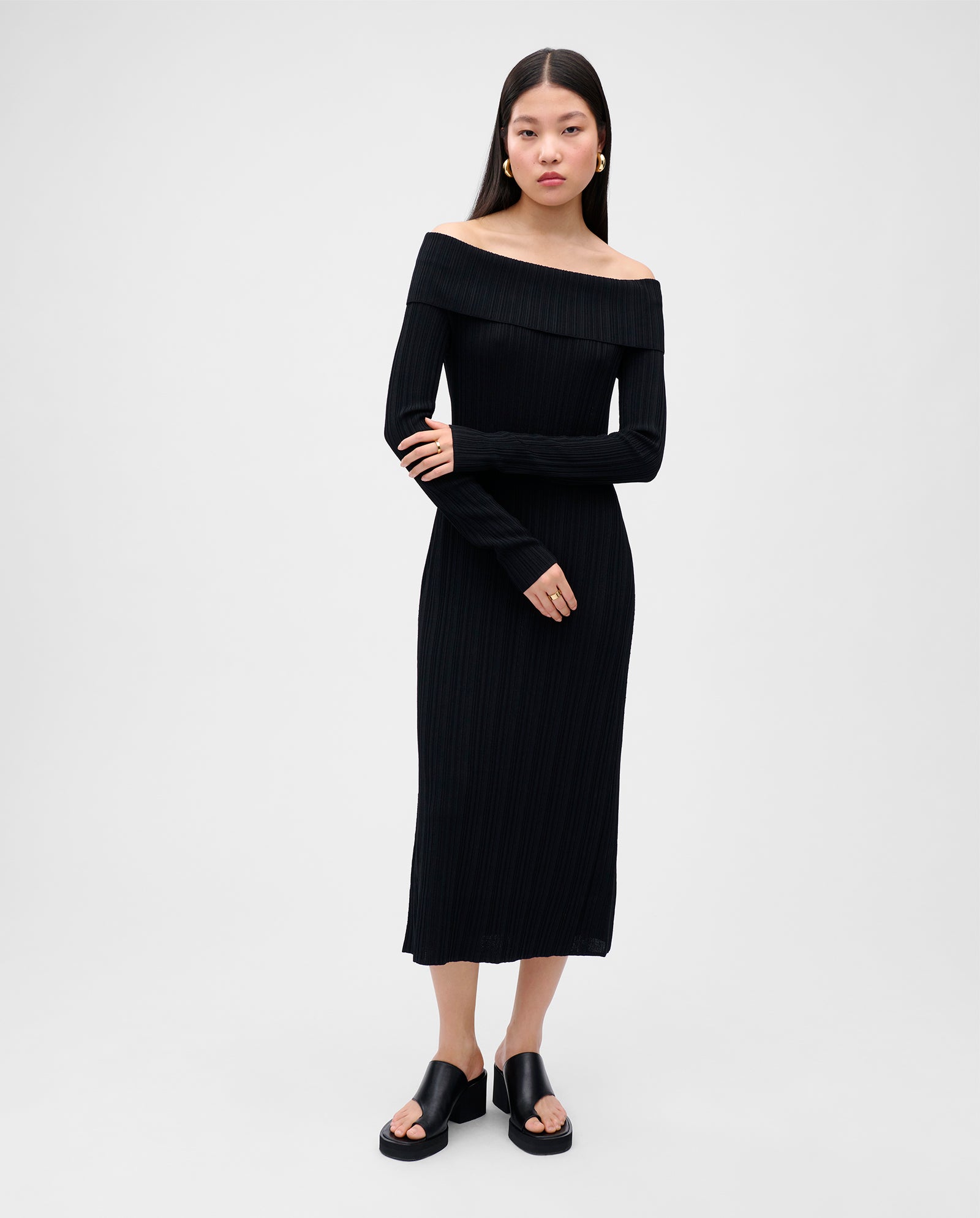 Woman in a black, figure-hugging IVY OAK KANDICE Dress with an off-the-shoulder carmen neckline and long sleeves, styled with black platform sandals.