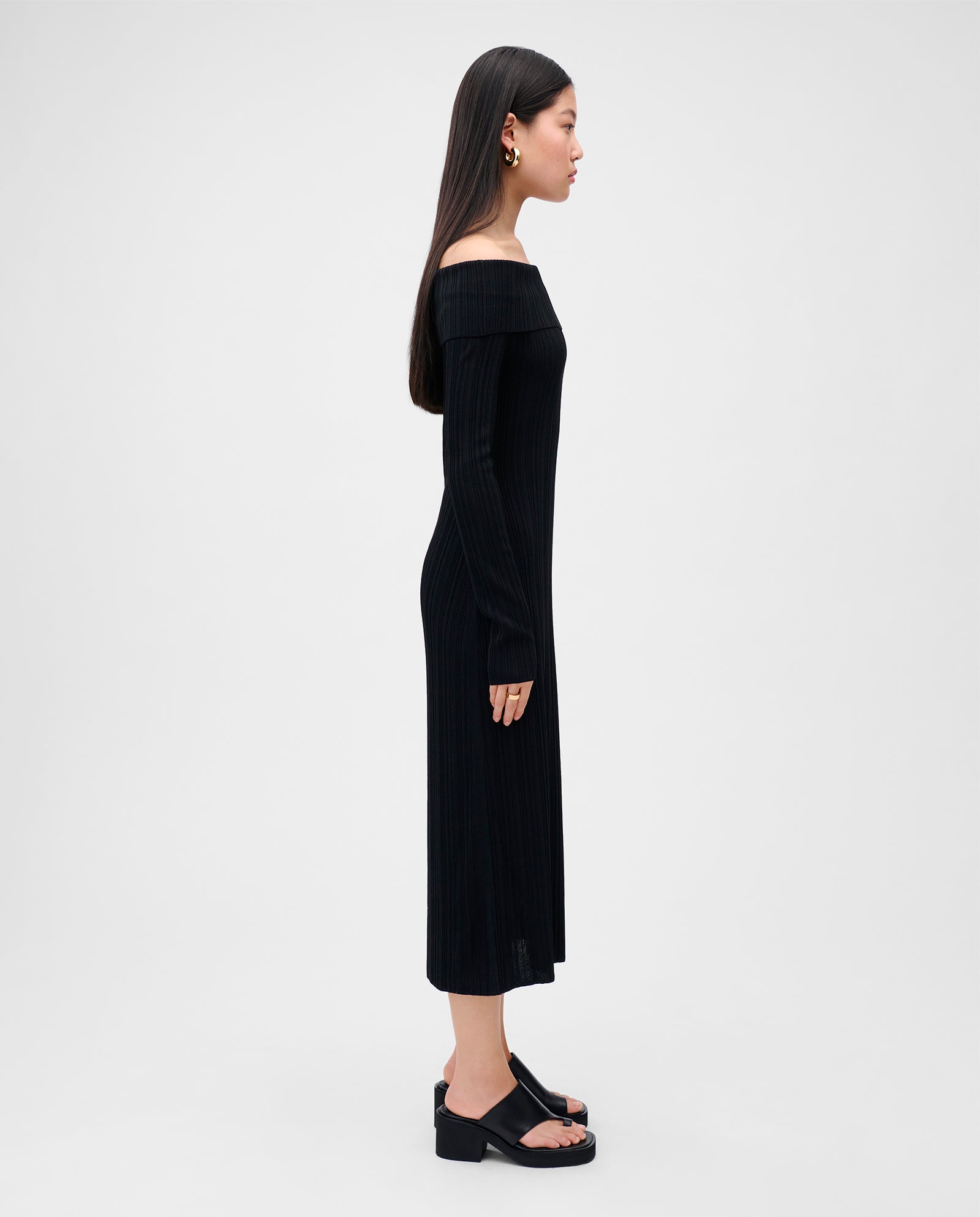 Woman in a black, figure-hugging KANDICE Dress by IVY OAK with a carmen neckline and long sleeves, standing in profile against a white background.