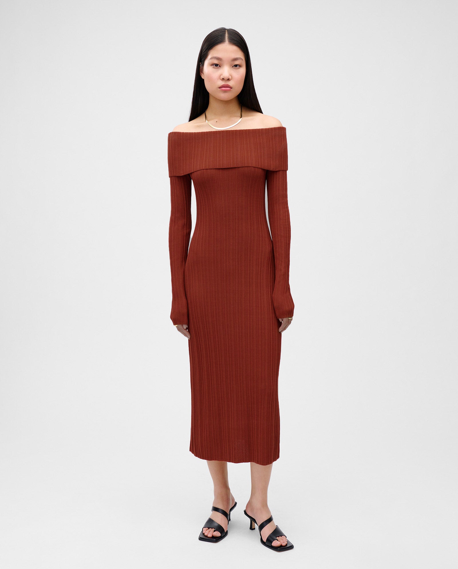 A woman stands neutrally in an IVY OAK KANDICE Dress—long, figure-hugging, rust-colored knit with an off-the-shoulder carmen neckline—paired with black heeled sandals.