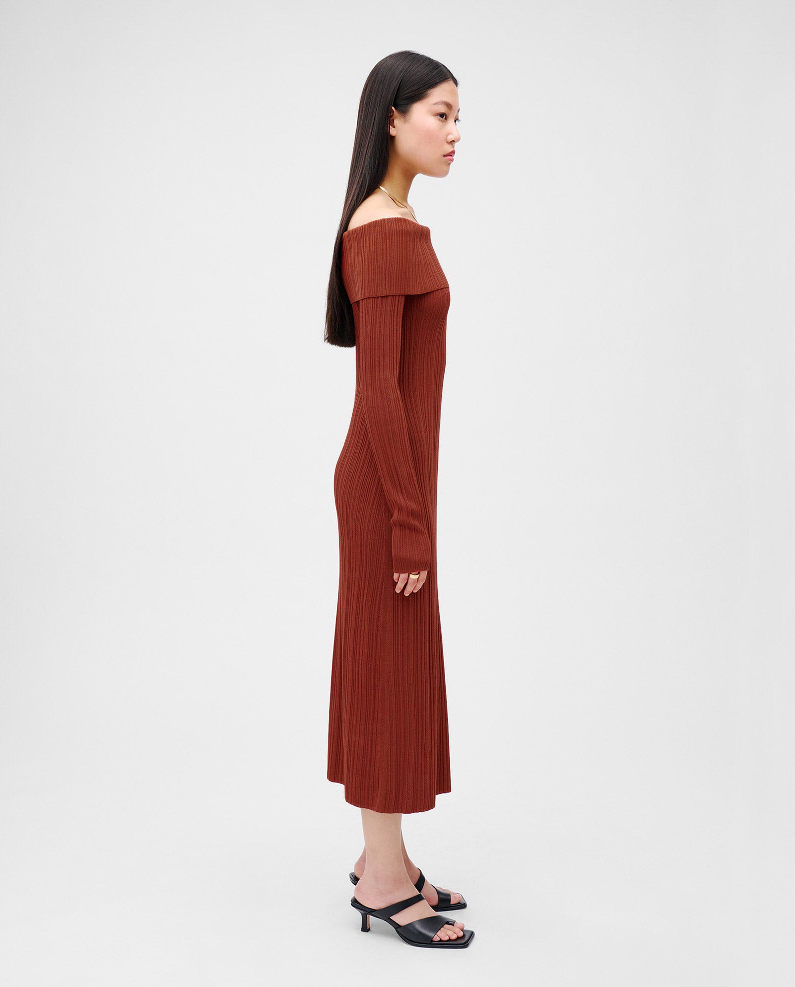 Woman in a rust-colored, off-shoulder IVY OAK KANDICE Dress with a carmen neckline and black heels stands in profile against a plain background. The figure-hugging silhouette enhances her shape.