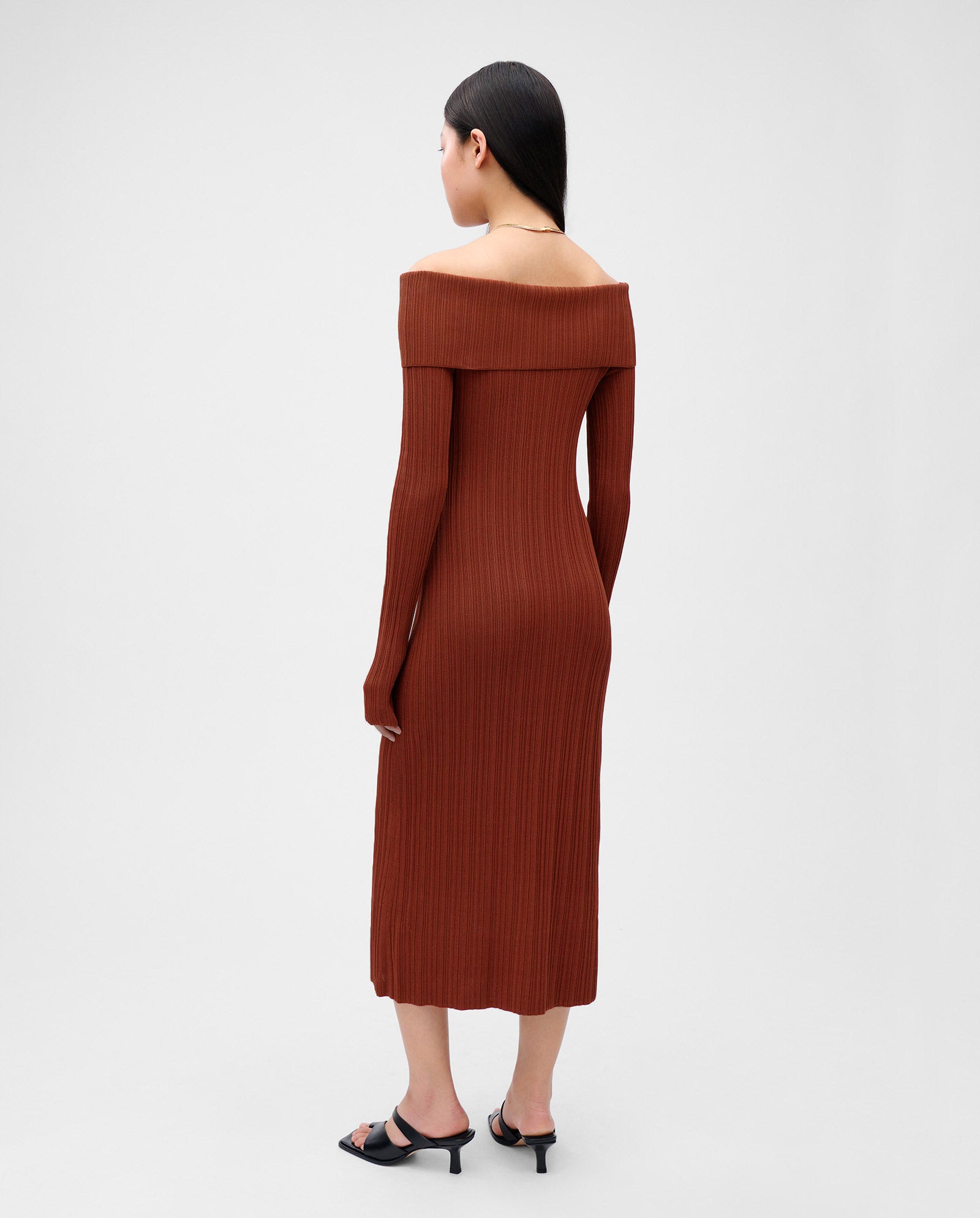 A woman, standing with her back to the camera, wears the IVY OAK KANDICE Dress—an off-shoulder, figure-hugging brown knit with long sleeves and ribbed fabric—paired with black heels.