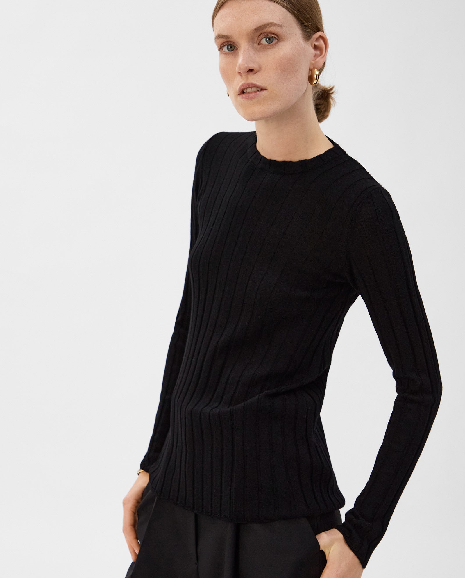 A woman poses against a white background wearing the IVY OAK KACY ANN Top, a fitted black Merino rib pullover paired with black pants—an ideal look for the transitional season.