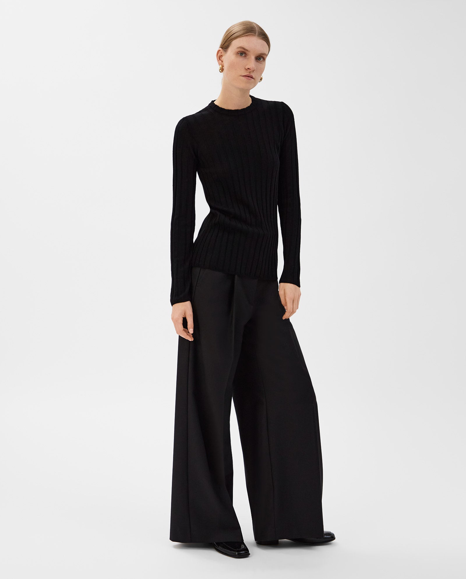 A woman wears the IVY OAK KACY ANN Top, a fitted black Merino rib pullover, paired with wide-leg black trousers against a white background—ideal for the transitional season.