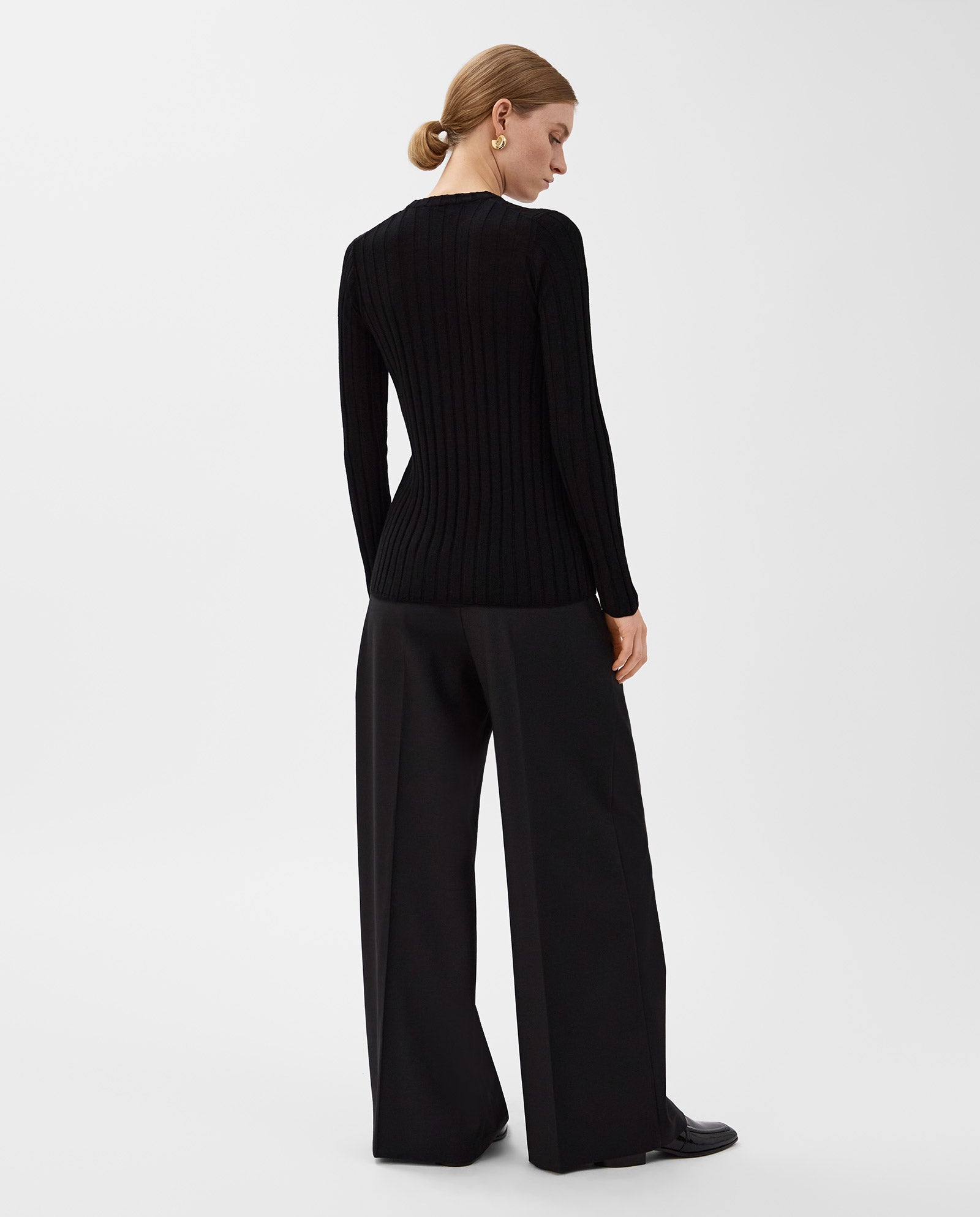 A woman in black wide-leg pants and the fitted KACY ANN Top by IVY OAK stands facing away against a plain background, embodying effortless style for the transitional season.