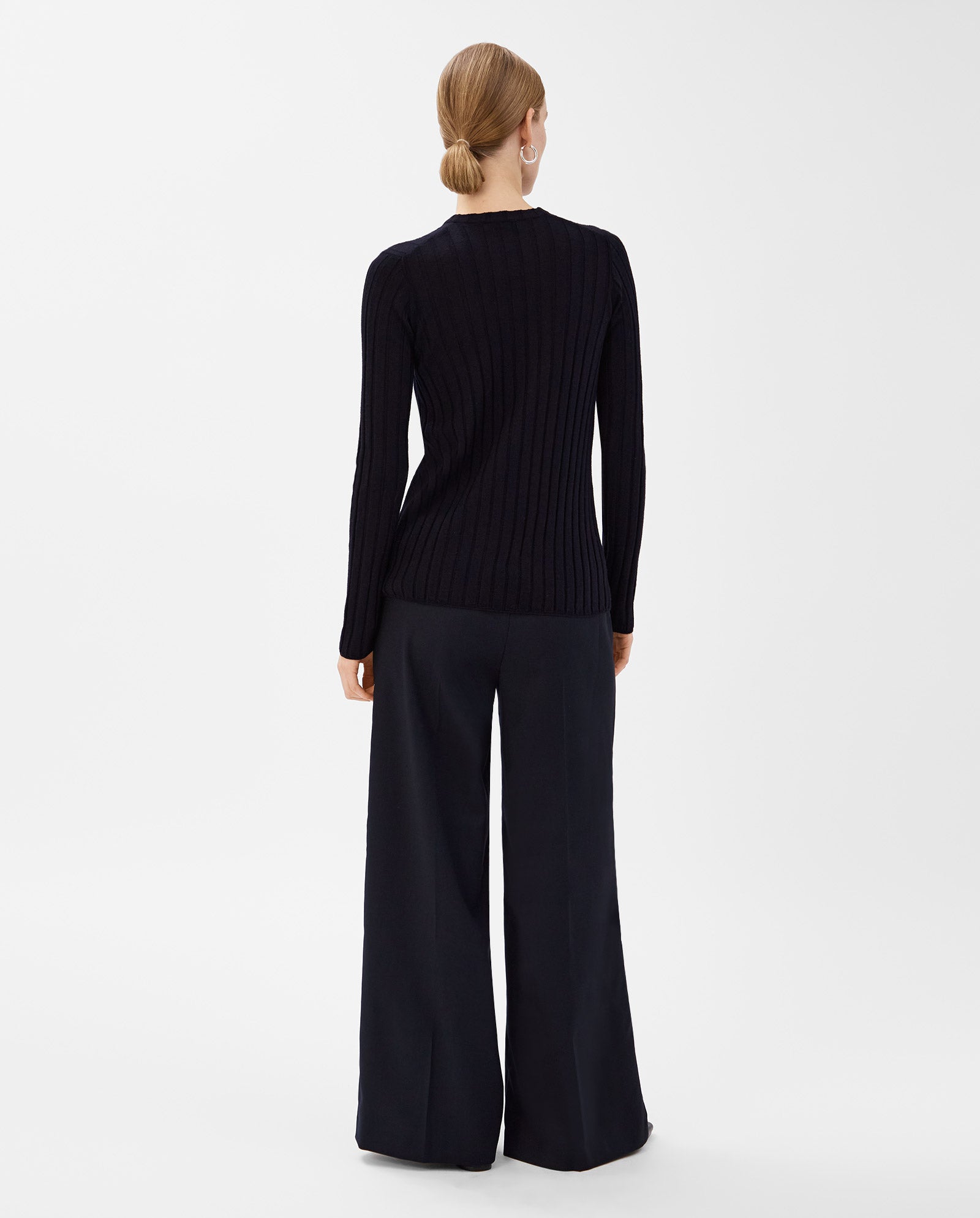 A woman stands with her back to the camera, wearing the IVY OAK KACY ANN Top and wide-leg black pants—an effortlessly chic look for the transitional season.