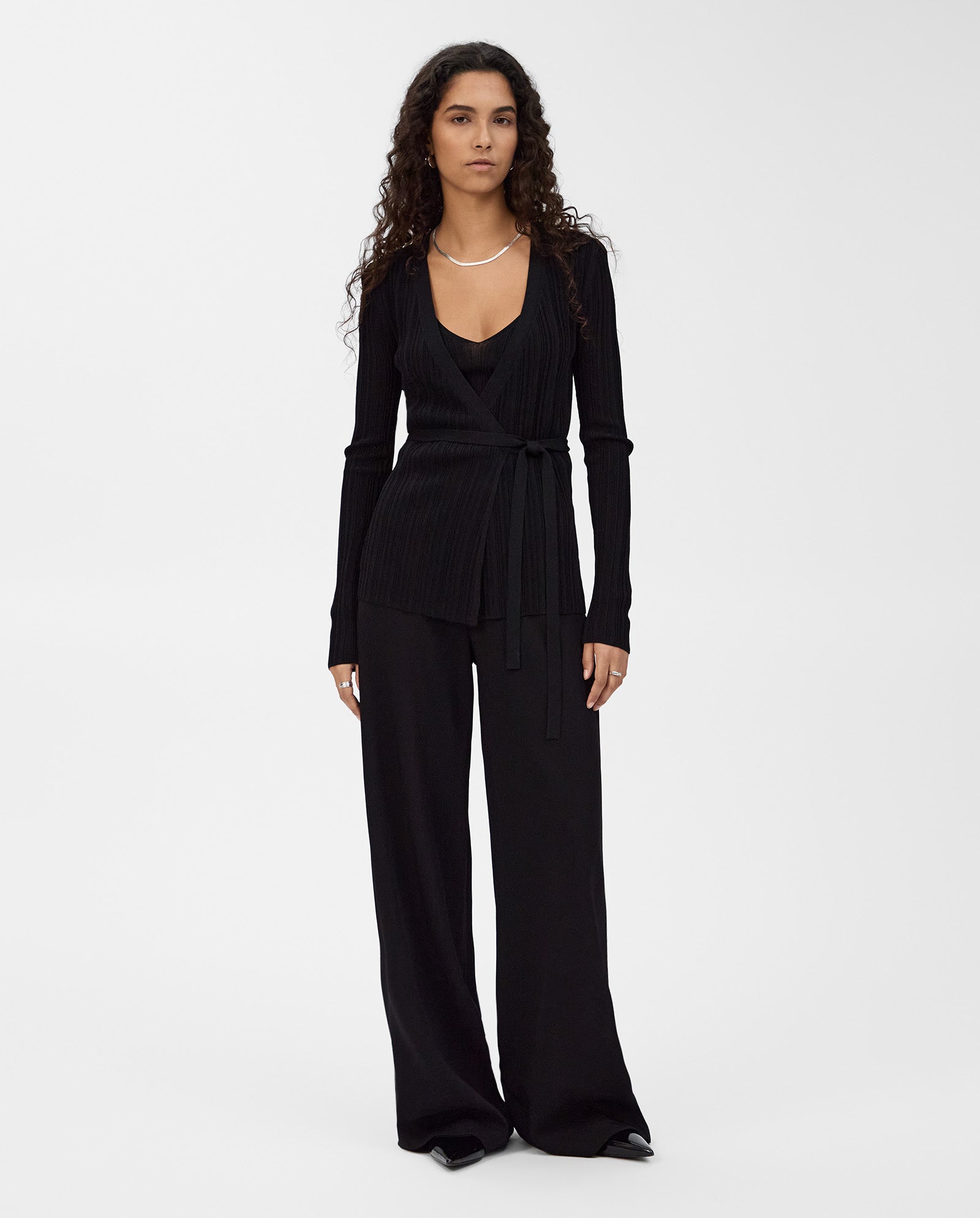 A woman with long curly hair wears the IVY OAK KIERA Cardigan and wide-leg black pants, standing against a white background.