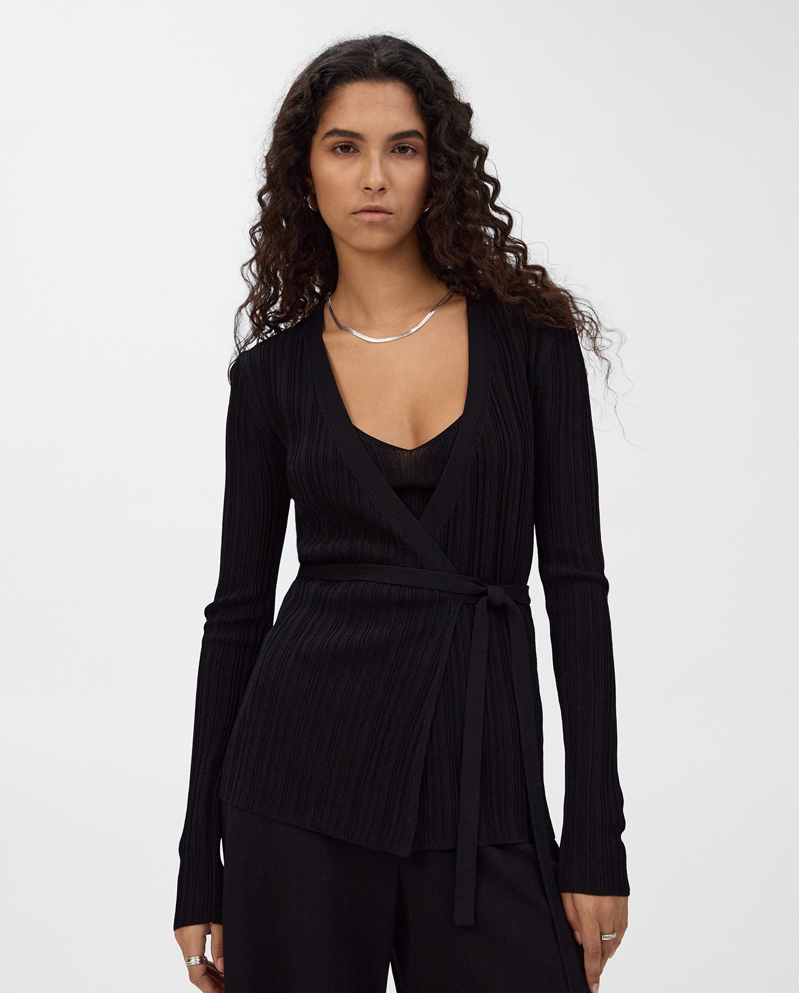 A woman with long curly hair wears the IVY OAK KIERA Cardigan, a black viscose wrap blouse with elegant sleeves, layered over a black shirt and black pants.