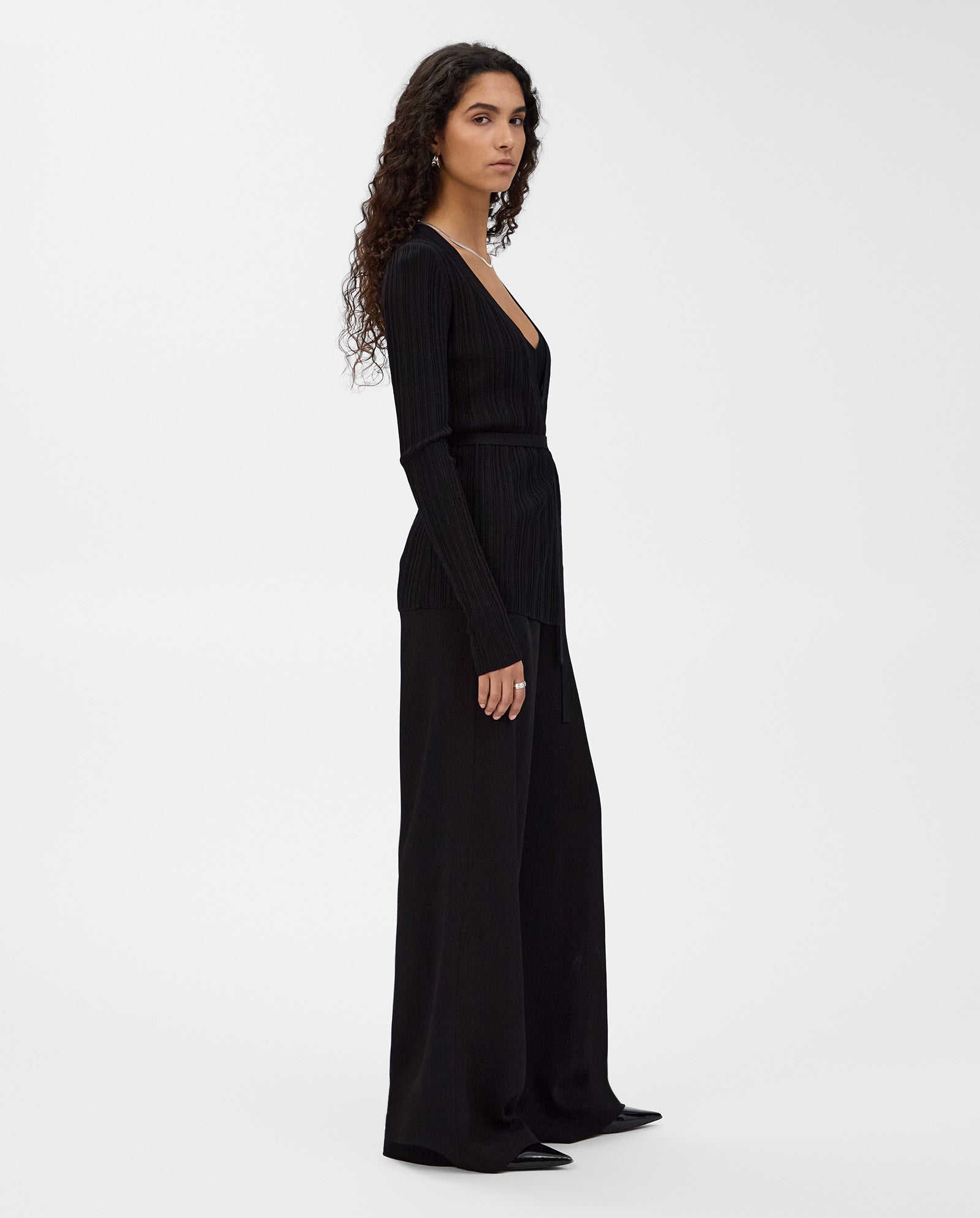 Woman with long curly hair wearing the IVY OAK KIERA Cardigan, styled with black pants and shoes, standing sideways.