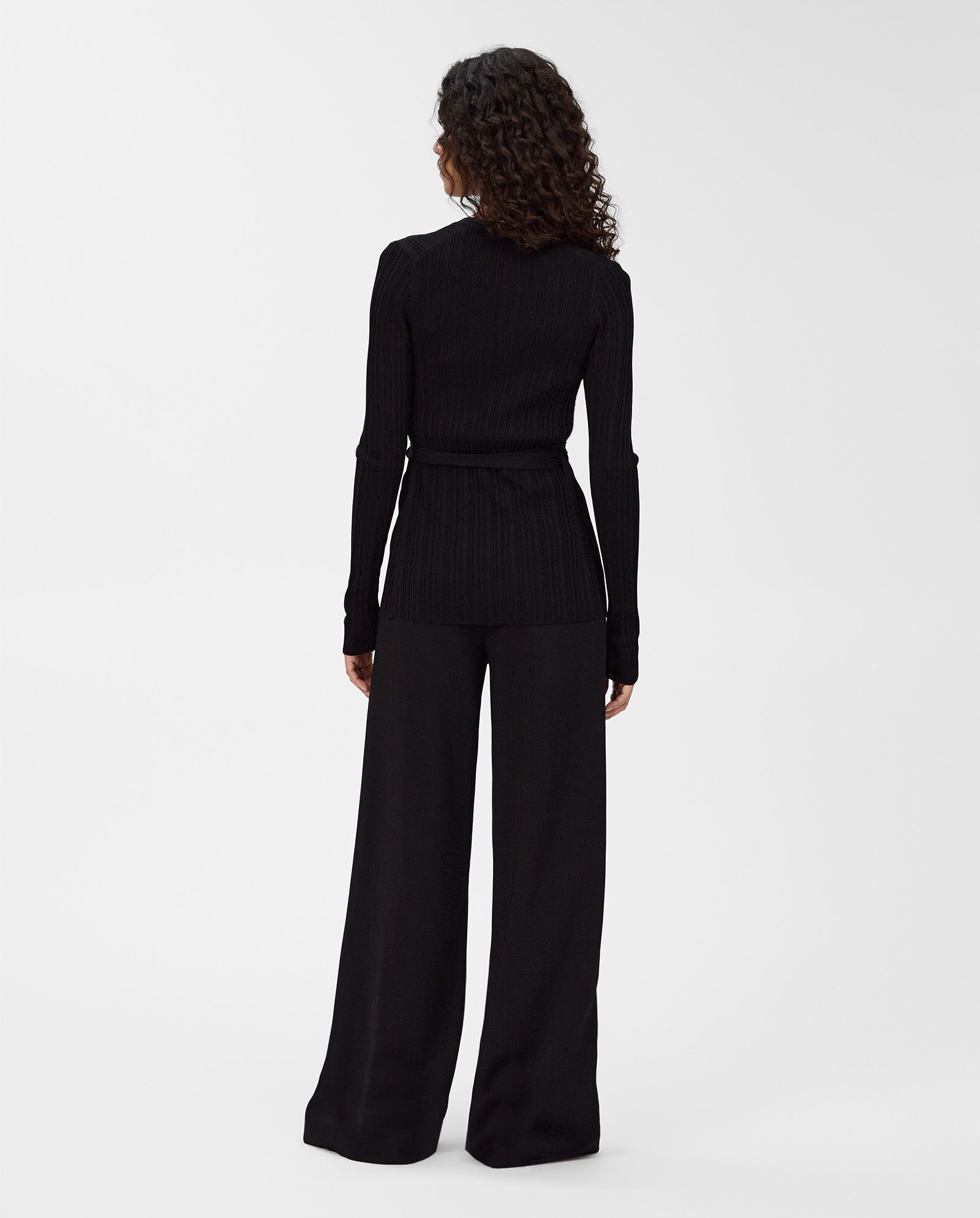 A person with curly hair, facing away from the camera, wears the IVY OAK KIERA Cardigan in black with elegant sleeves and pairs it with wide-leg pants.