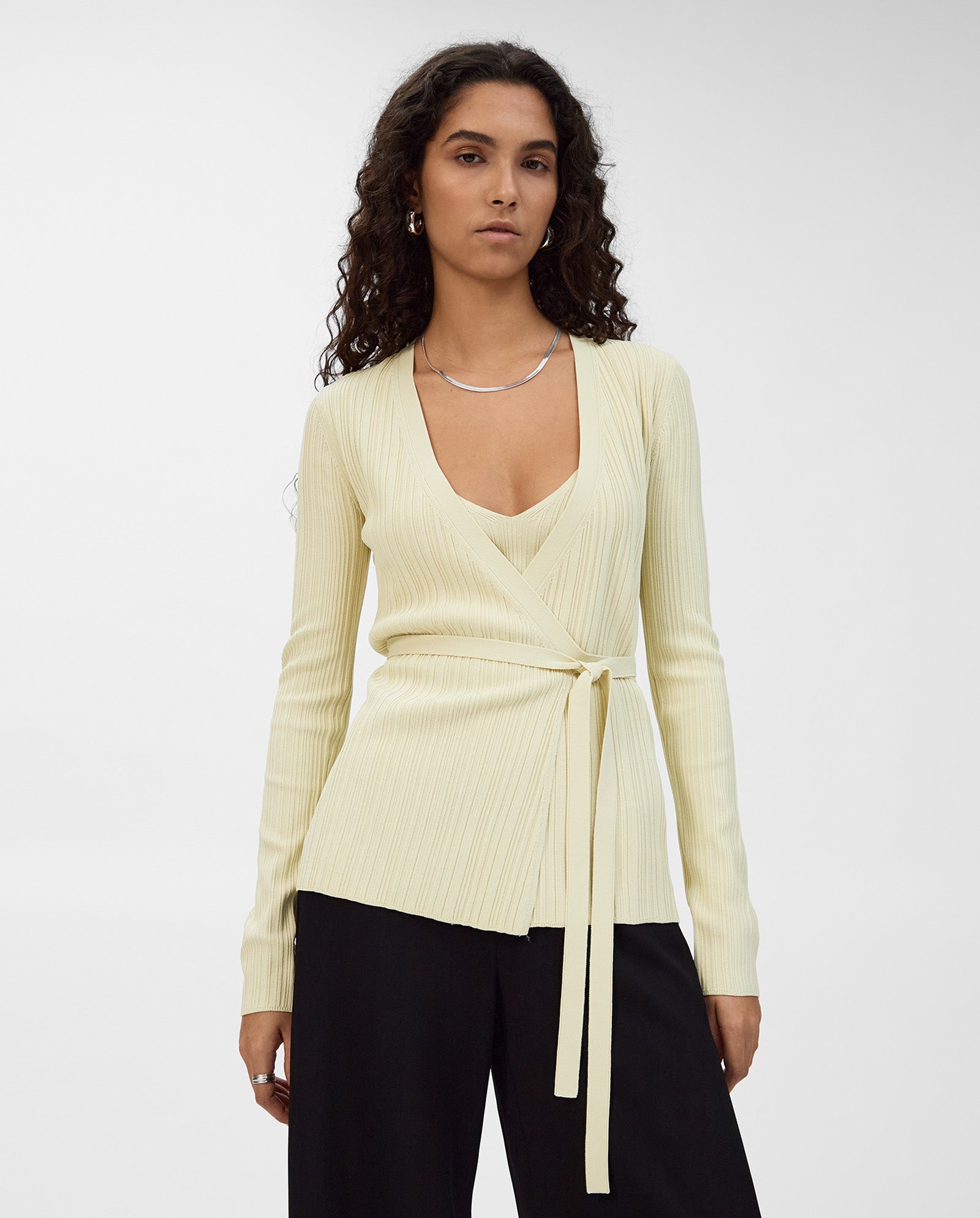 A woman stands against a plain background wearing the IVY OAK KIERA Cardigan, a light yellow viscose wrap blouse with waist tie, elegant sleeves, and black pants.