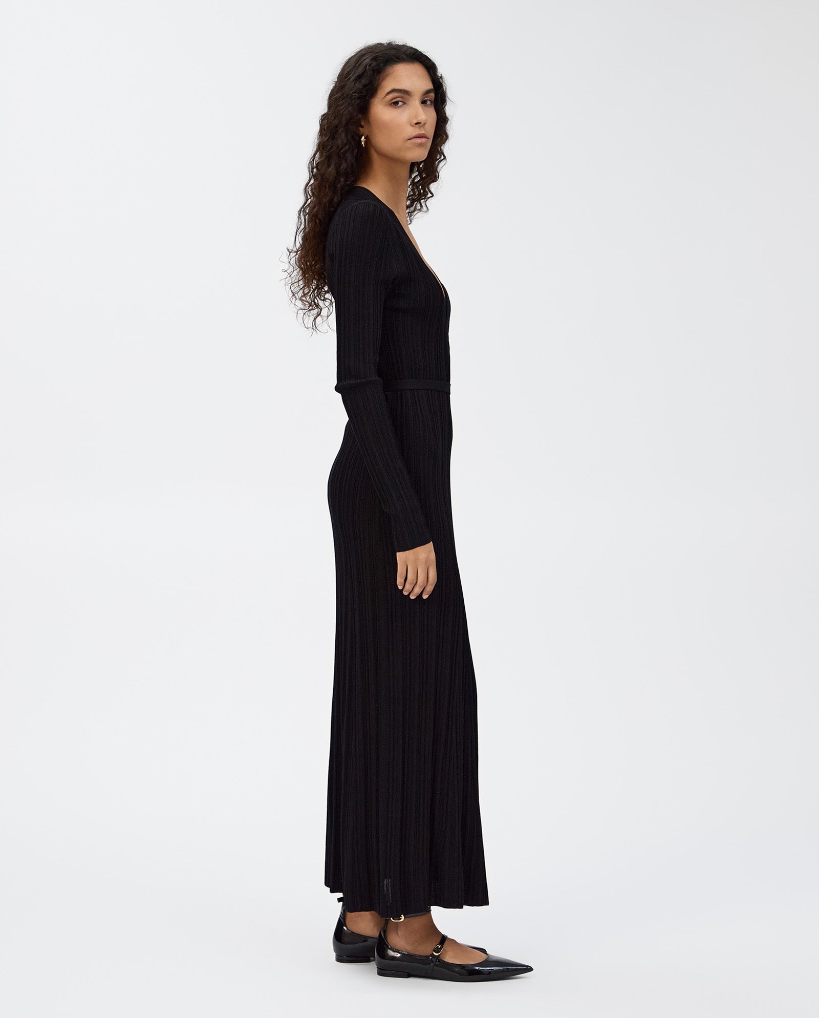 A woman with long curly hair stands sideways, wearing the IVY OAK KLOE Dress—a black viscose wrap dress with stretchy sleeves—paired with black flats.