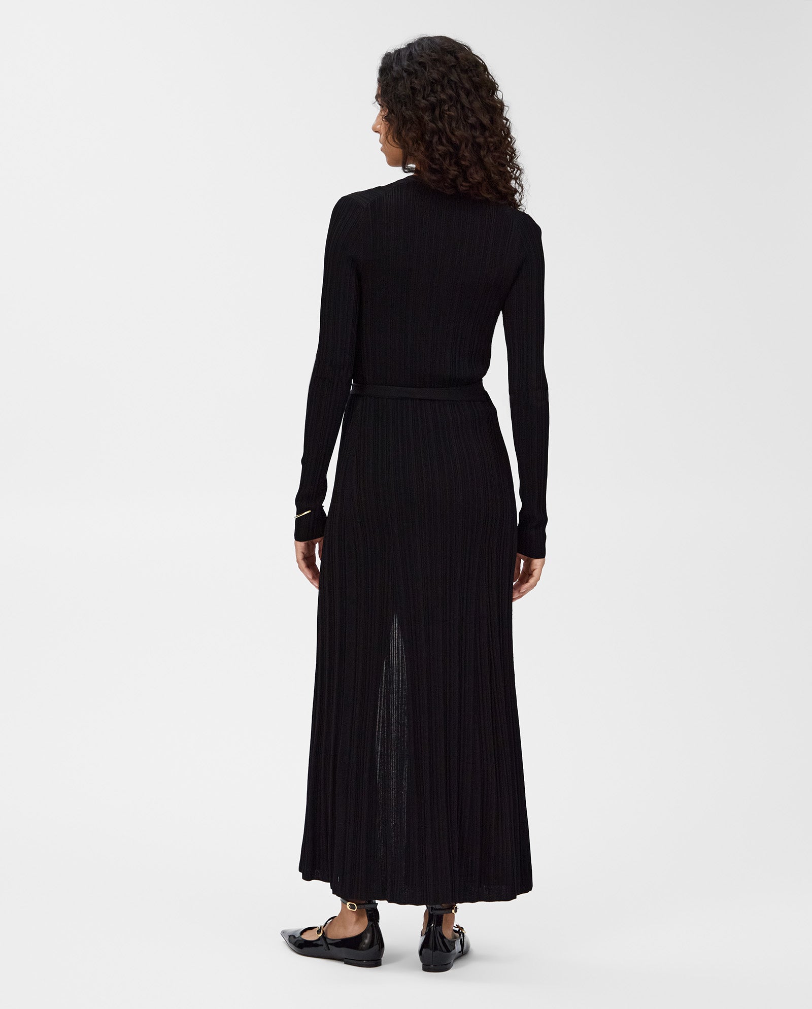 A woman stands with her back to the camera, wearing the IVY OAK KLOE Dress—a long-sleeved, ankle-length black viscose wrap dress—paired with black shoes.