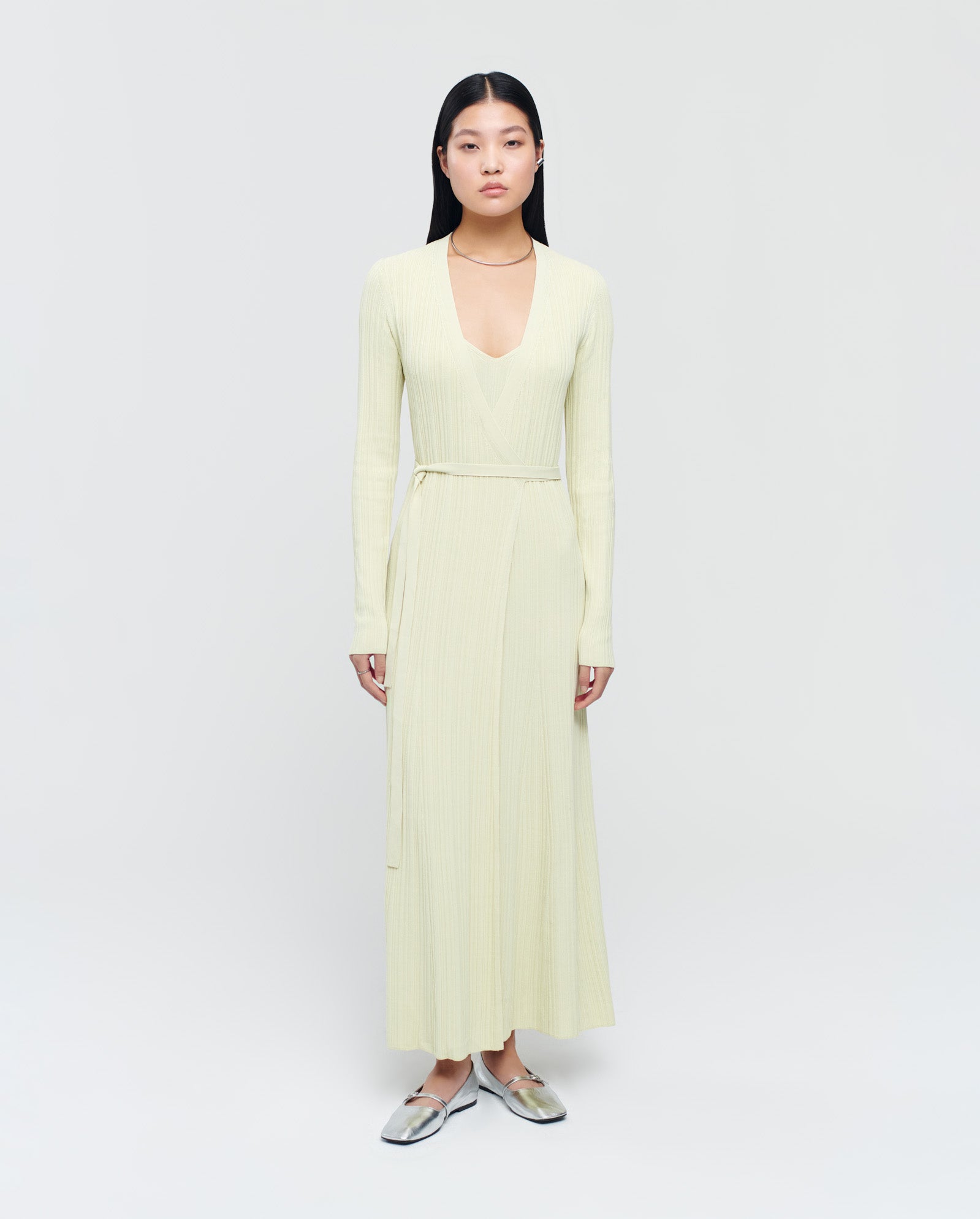 A woman wears the IVY OAK KLOE Dress, a long pale yellow viscose wrap dress with stretchy sleeves, paired with silver shoes against a plain background.