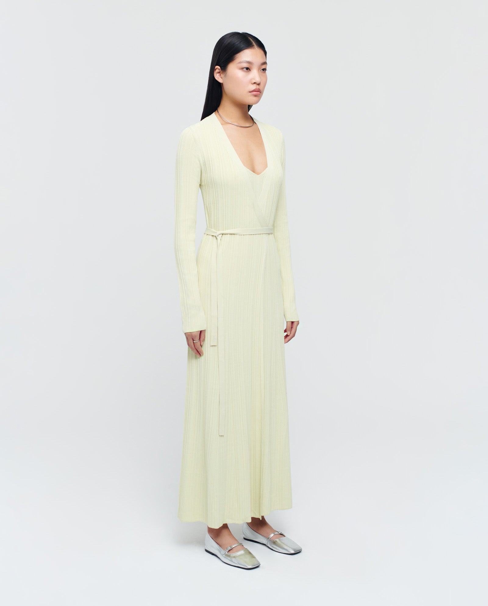 A woman wearing the IVY OAK KLOE Dress, a long light yellow wrap dress with stretchy sleeves, silver flats, and straight black hair stands against a plain background.