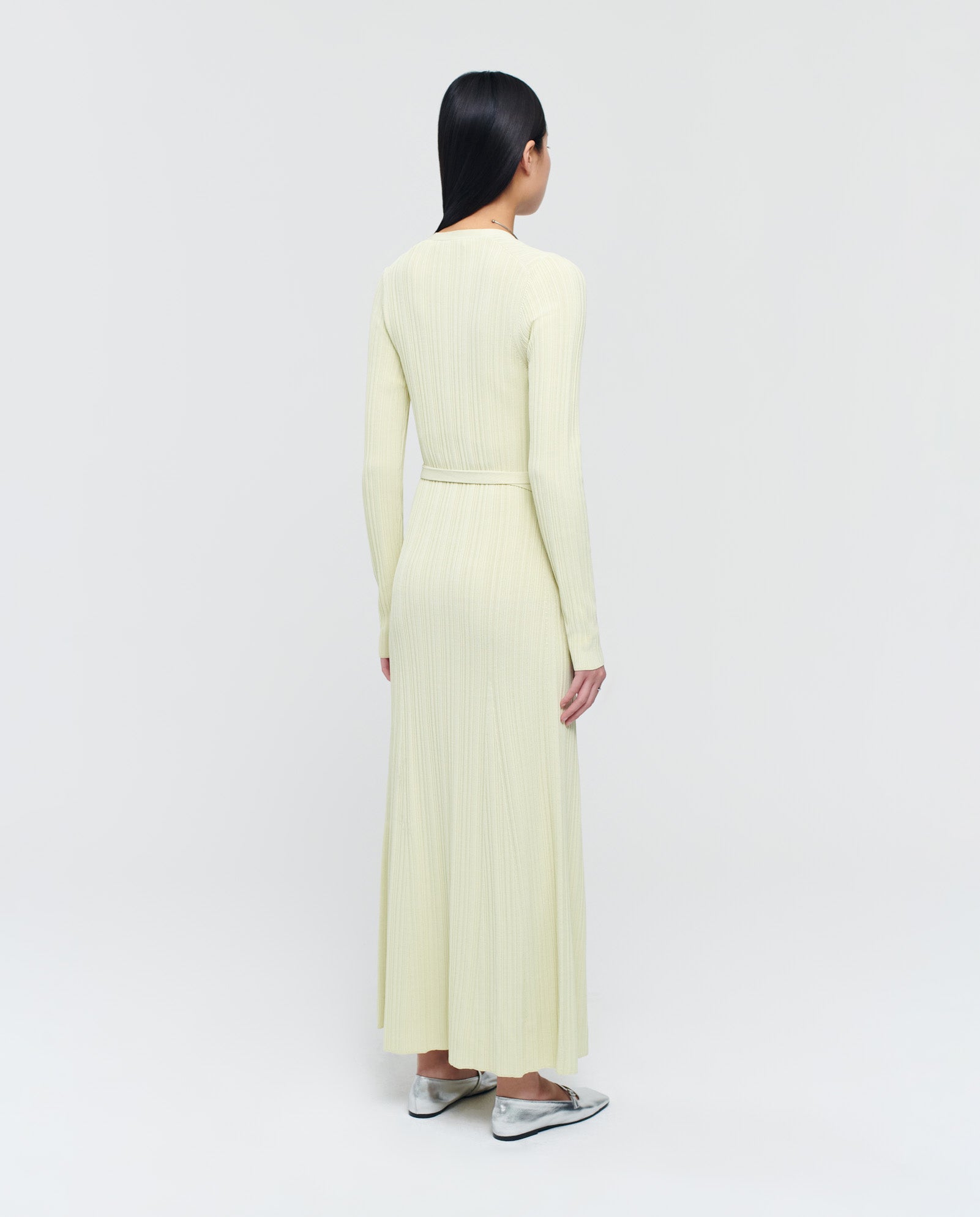 A woman stands with her back turned, wearing the IVY OAK KLOE Dress—a pale yellow viscose wrap dress with stretchy sleeves—and silver flats, set against a plain background.