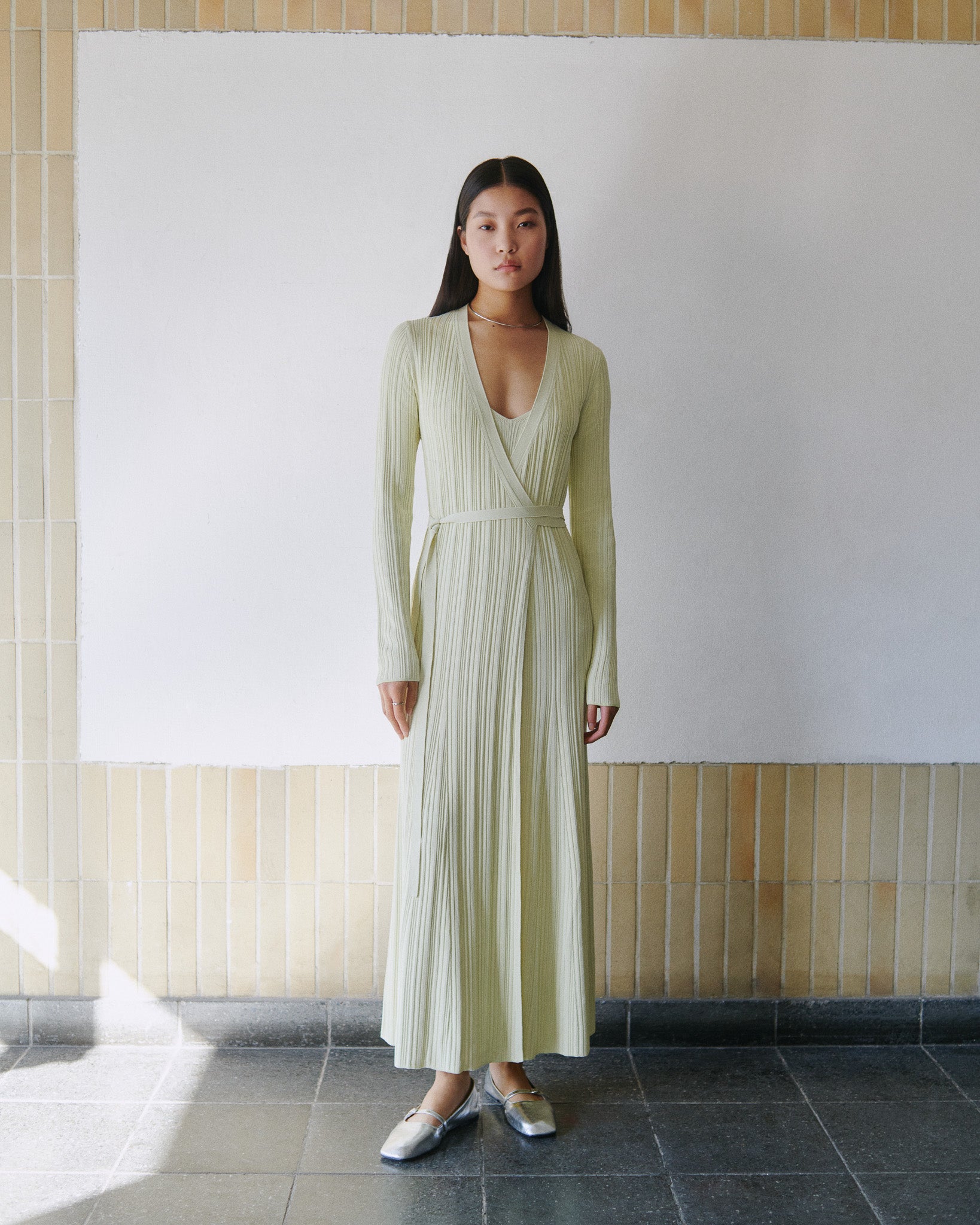 A woman models the IVY OAK KLOE Dress, a long light green viscose wrap dress with stretchy sleeves and a graceful flared silhouette, standing indoors against a tiled wall and white backdrop.