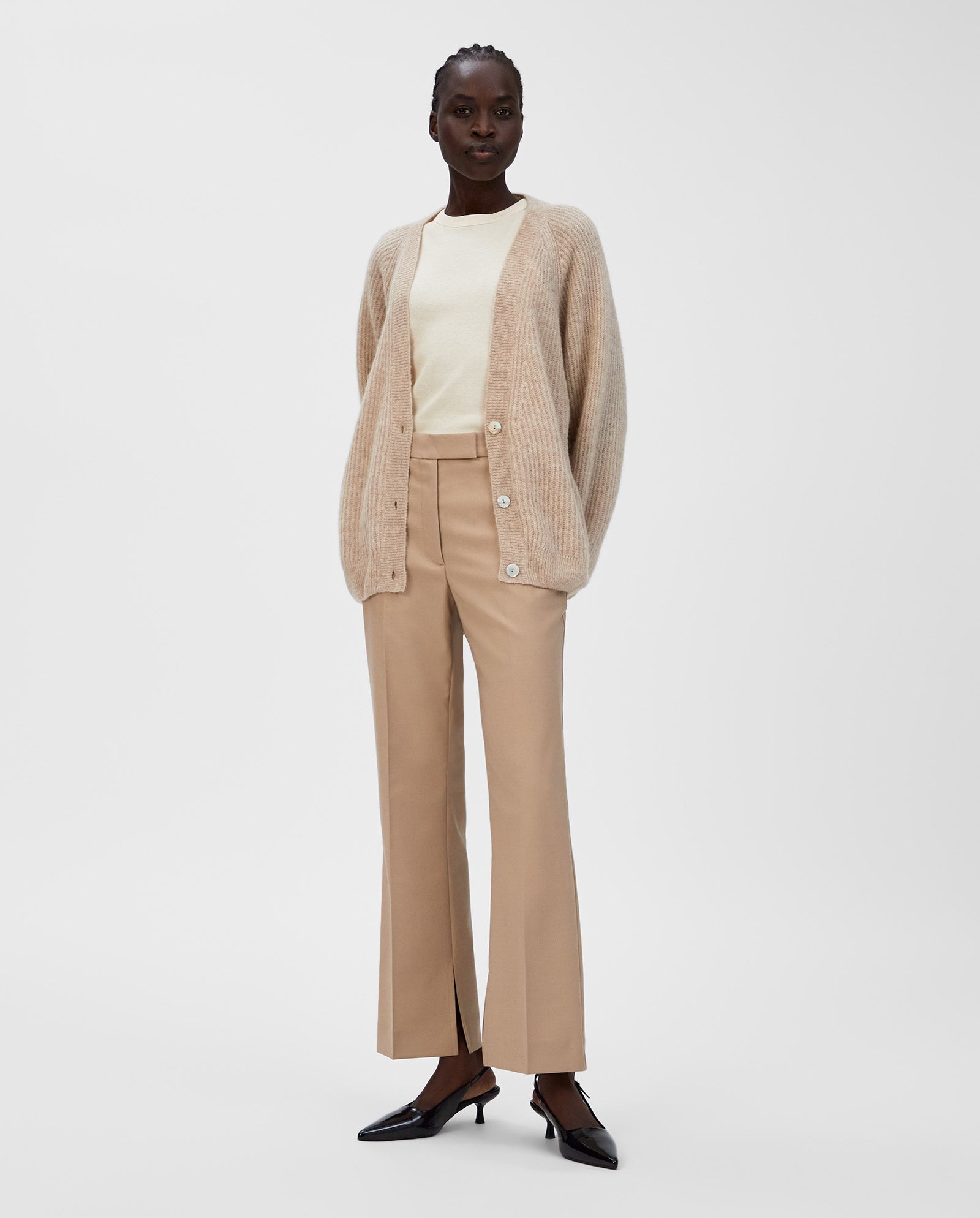 A woman stands against a white background wearing the IVY OAK KEONA ANN Cardigan, an alpaca blend beige piece with a deep neckline, paired with beige pants and black heels.