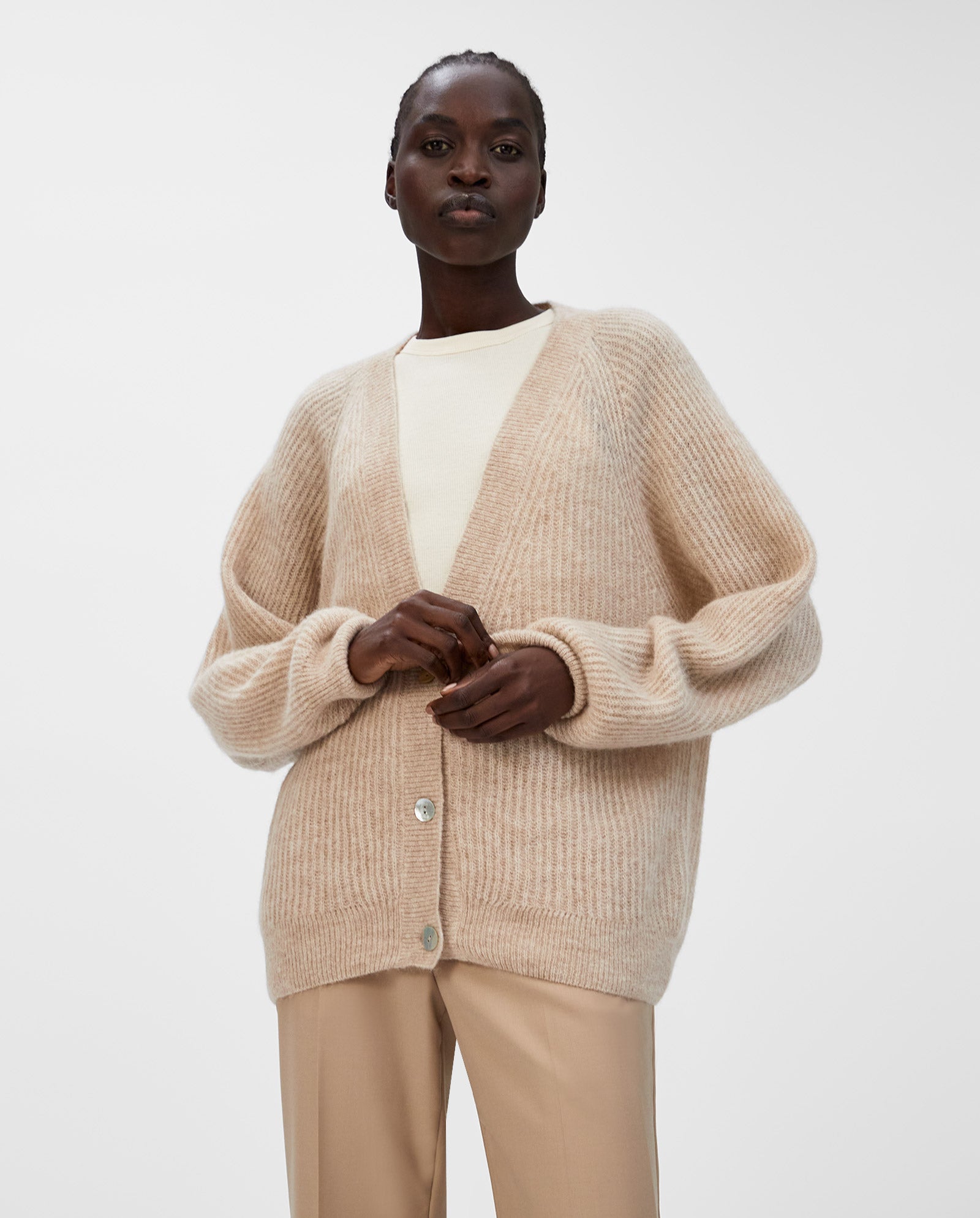 A person wears the IVY OAK KEONA ANN Cardigan, an alpaca blend beige piece with a deep neckline, over a cream top and tan pants against a plain background.