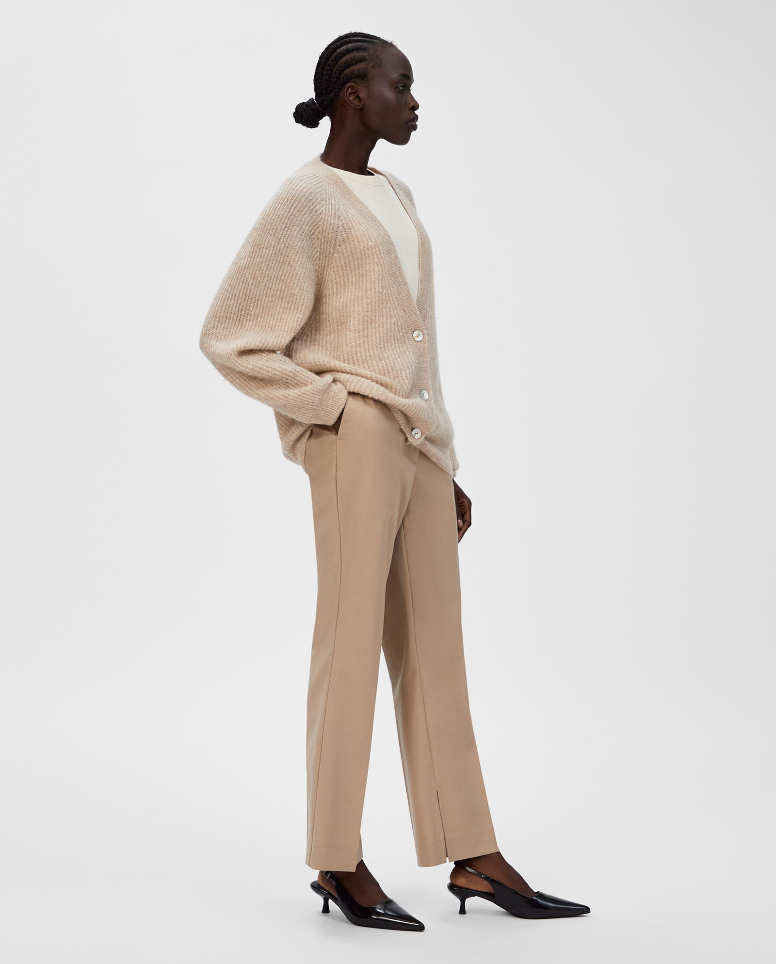 A woman in neutral tones stands sideways, wearing the IVY OAK KEONA ANN Cardigan—a beige alpaca blend with a deep neckline—paired with a cream top, tan pants, and black heels.