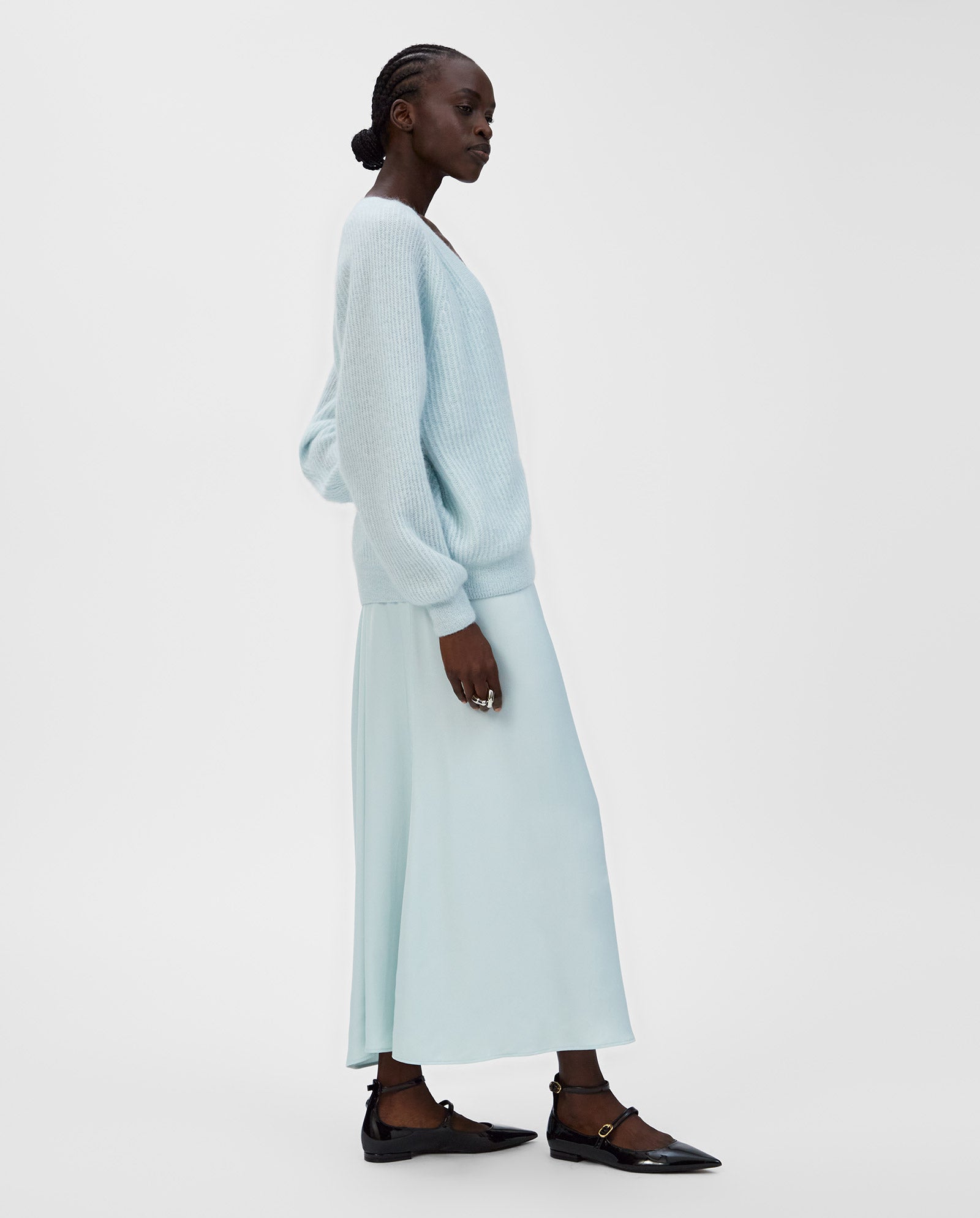 A woman wears the IVY OAK KEONA ANN cardigan with a deep neckline, styled with a light blue skirt and black flats, standing against a plain white background.