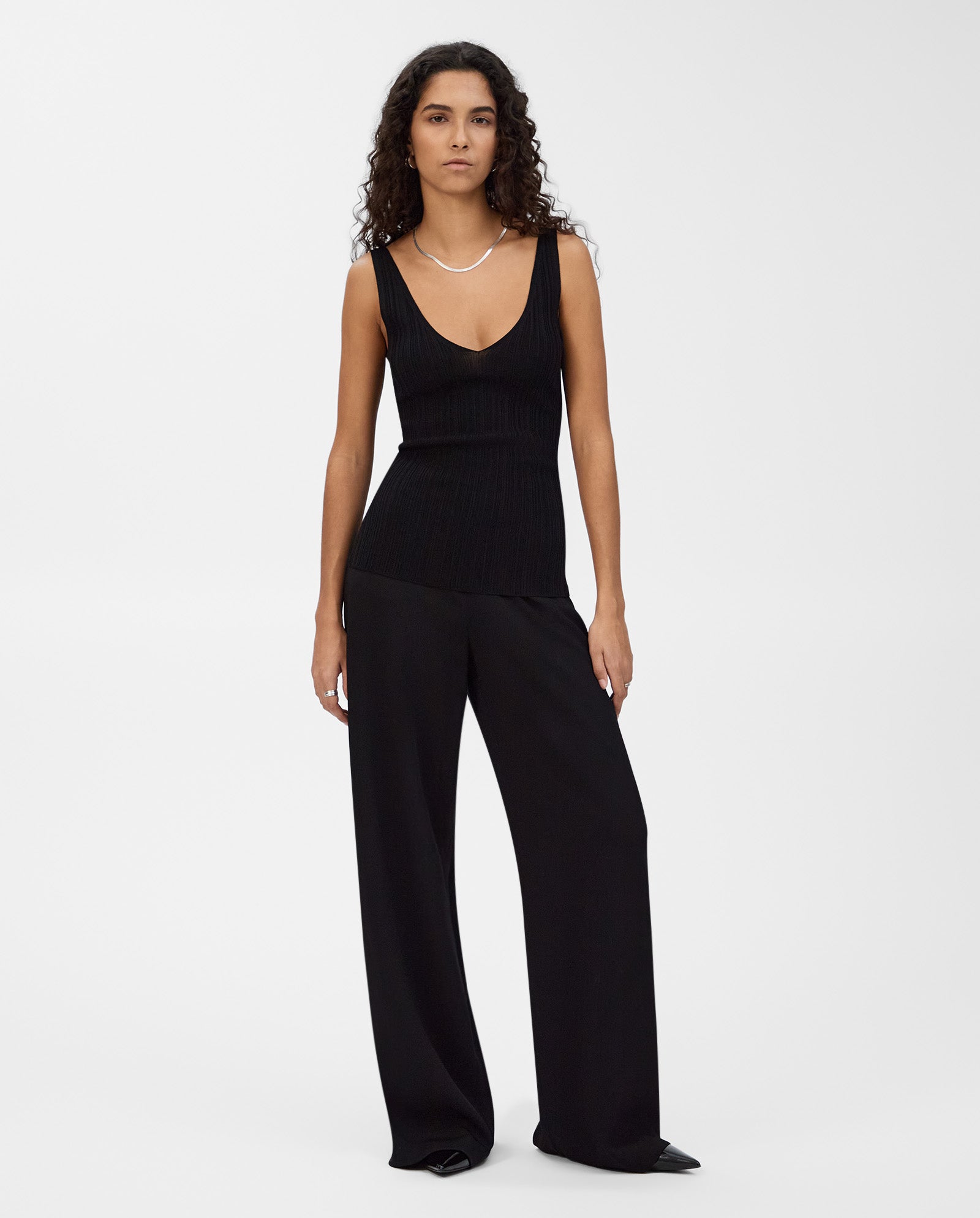 A woman wears the KYM Top by IVY OAK—a sleeveless, stretchy knit black top with a deep V-neckline—paired with wide-leg black pants, standing against a plain white background.