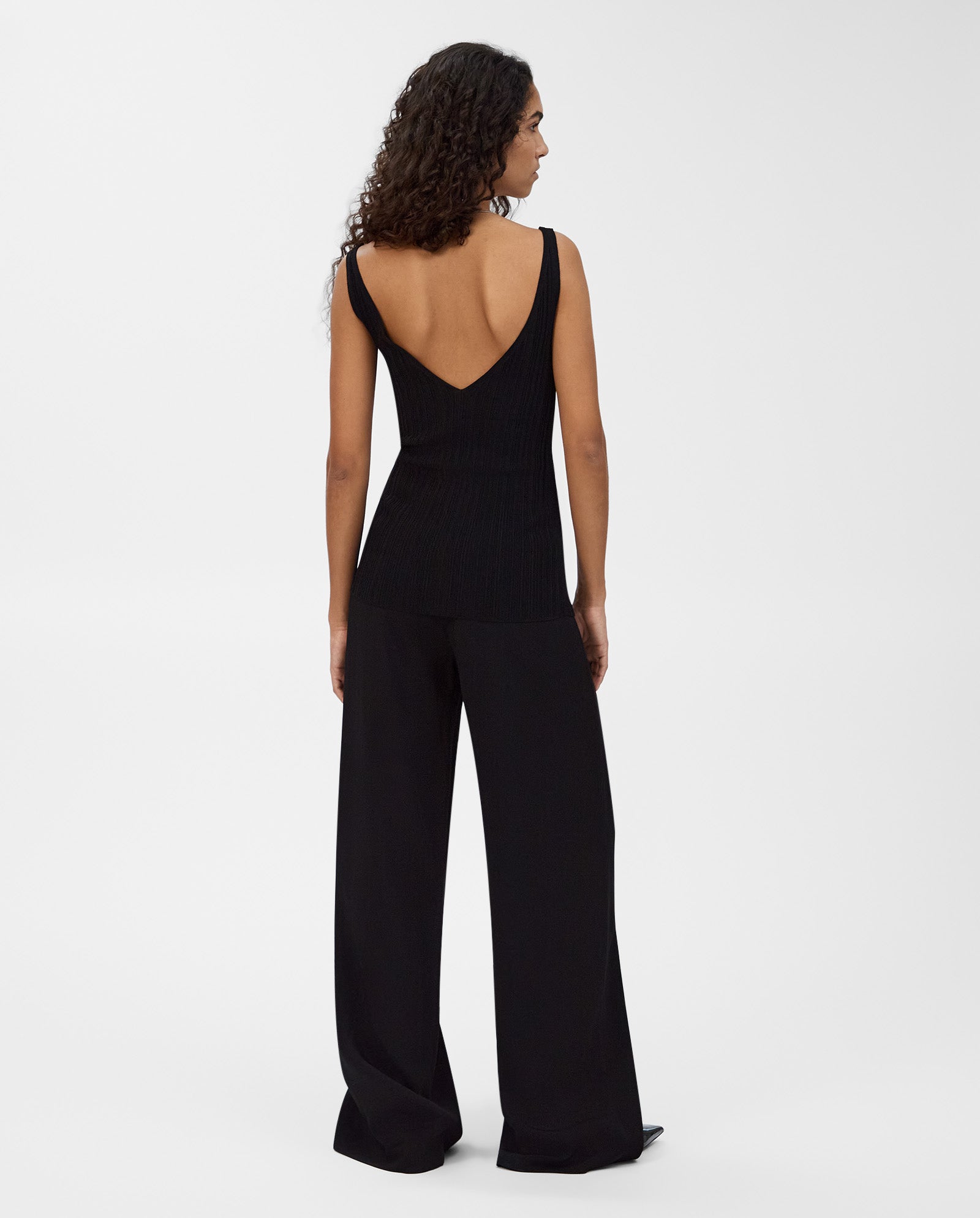 Woman with curly hair faces away wearing the IVY OAK KYM Top, a stretchy black knit with a deep V-back, paired with wide-leg black pants.