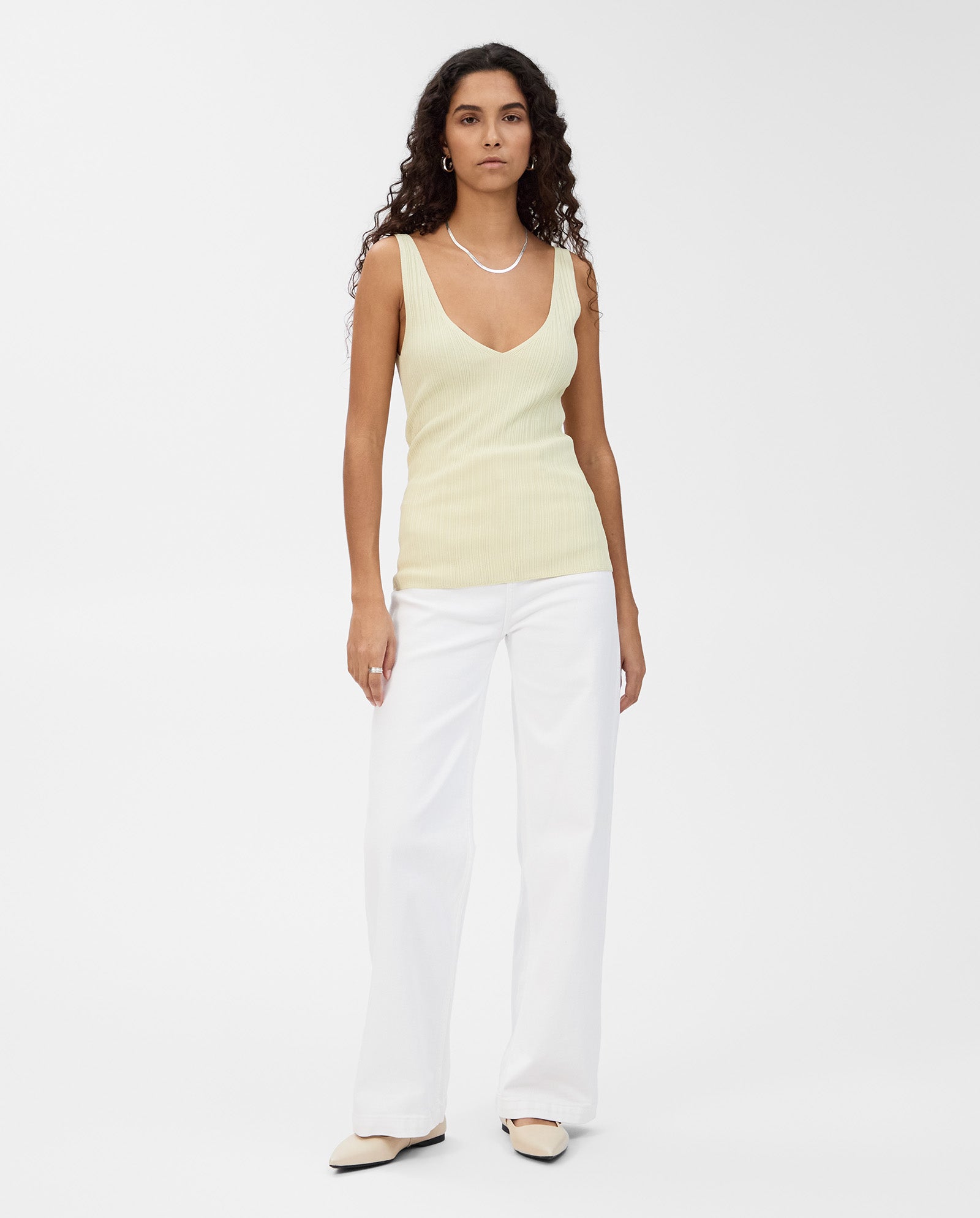 A woman wears the IVY OAK KYM Top, a yellow stretchy knit with a deep V-neckline, paired with white pants, cream shoes, and a necklace, standing against a white background.
