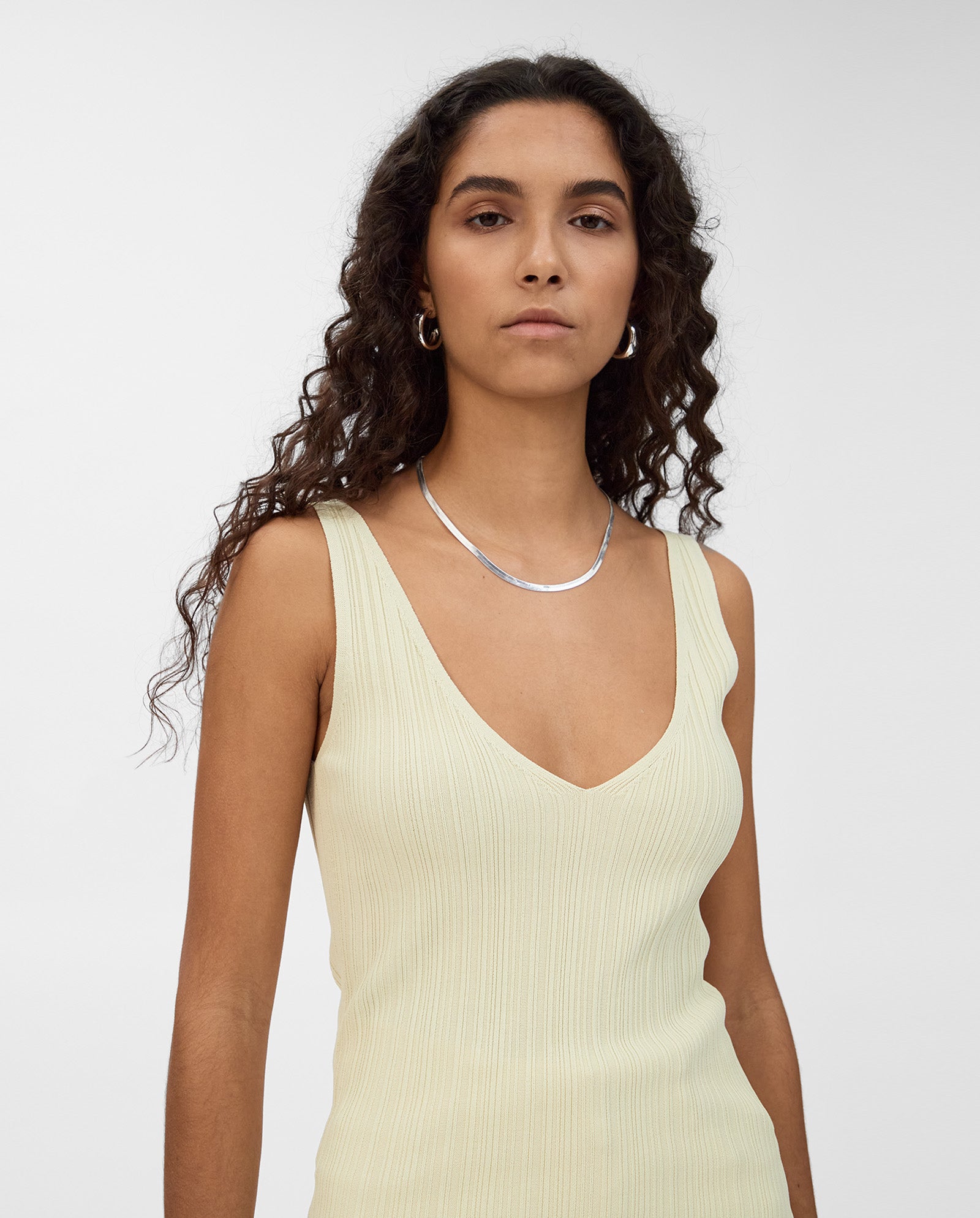 Woman with long curly hair wears the IVY OAK KYM Top, a sleeveless light yellow stretchy knit with a deep V-neckline, styled with silver jewelry against a plain background.