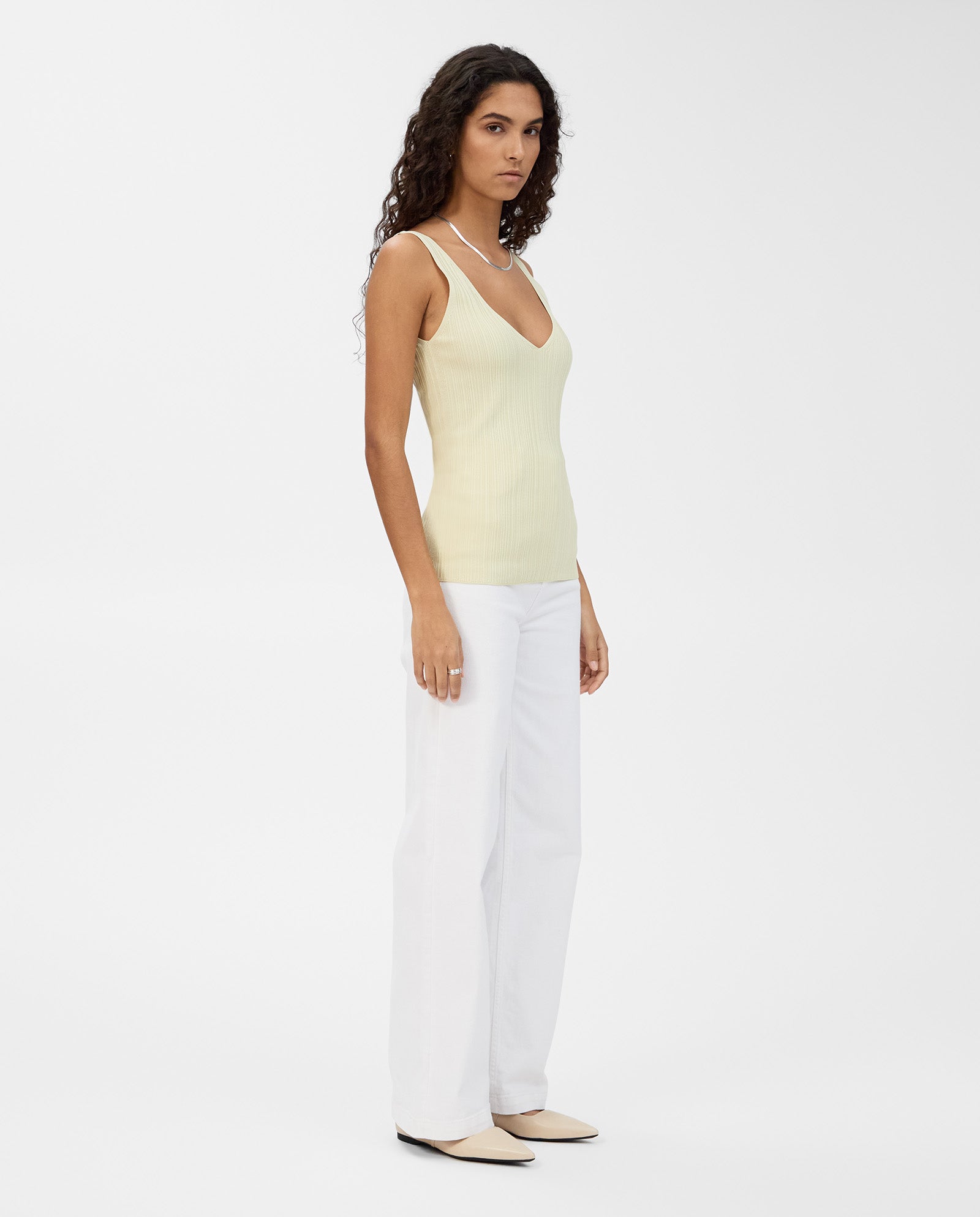 Woman in an IVY OAK KYM Top in pale yellow stretchy knit, paired with white pants and beige flats, stands against a plain white background.