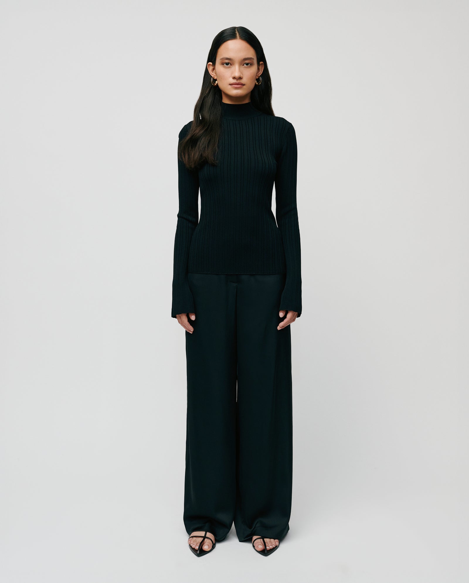 A woman wears the IVY OAK KYLIE Jumper, a black long-sleeve knitted viscose top with a stand-up collar, paired with wide-leg pants, standing against a plain white background.