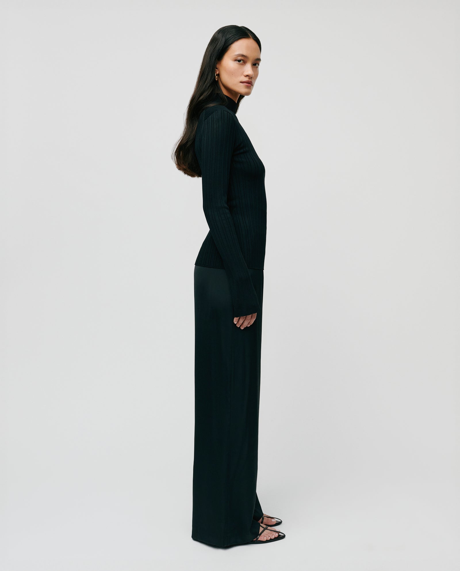 A woman stands in profile against a plain white background, wearing the IVY OAK KYLIE Jumper with long sleeves and a stand-up collar, paired with sandals.