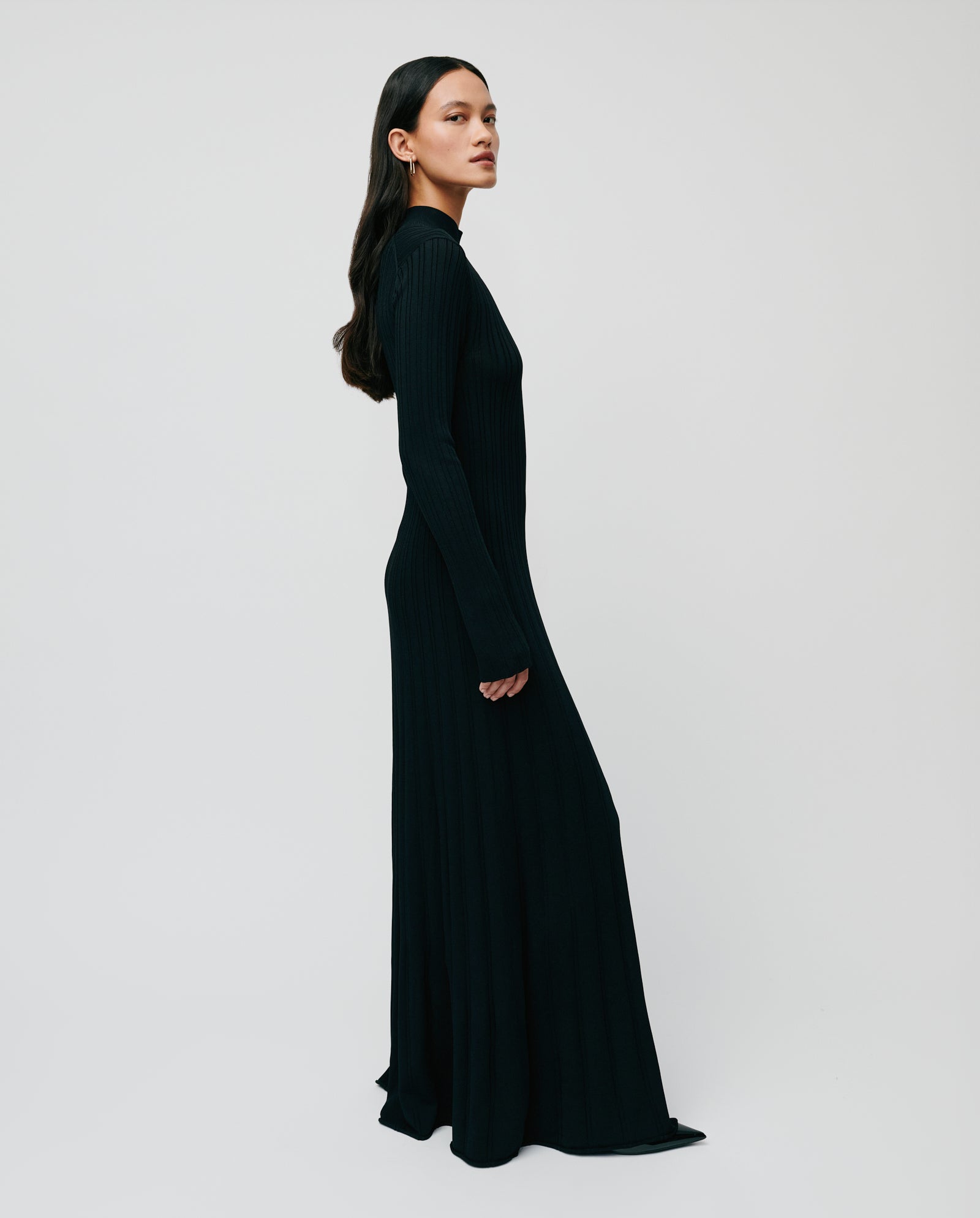 A woman stands sideways against a plain light background, wearing the IVY OAK KOURTNEY dress—a long-sleeved, floor-length viscose knit piece with a body-hugging fit.