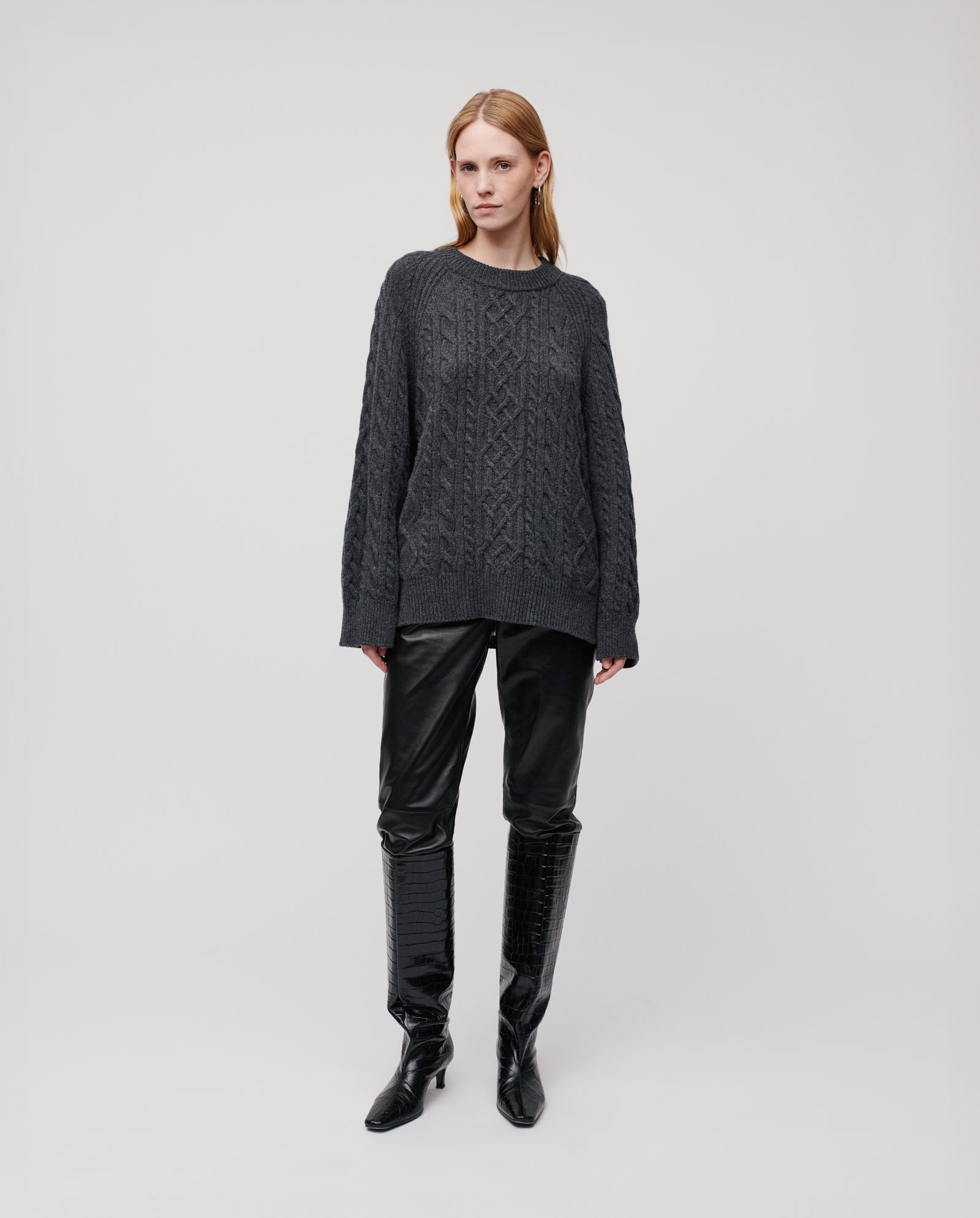 A woman stands in a relaxed pose, wearing the IVY OAK KLARINA ANN Jumper in gray, paired with black leather pants and knee-high black boots against a plain background.