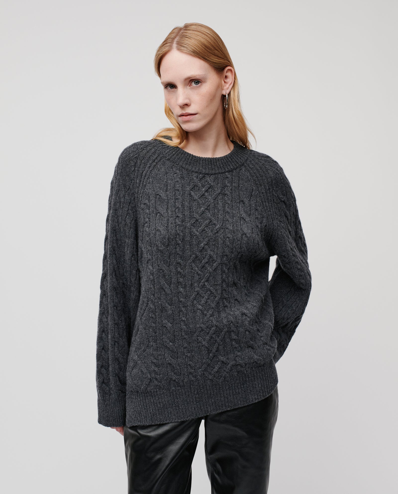 A woman wears the IVY OAK KLARINA ANN Jumper, a dark gray crewneck cable knit with a relaxed fit, paired with black pants, standing before a plain light background.