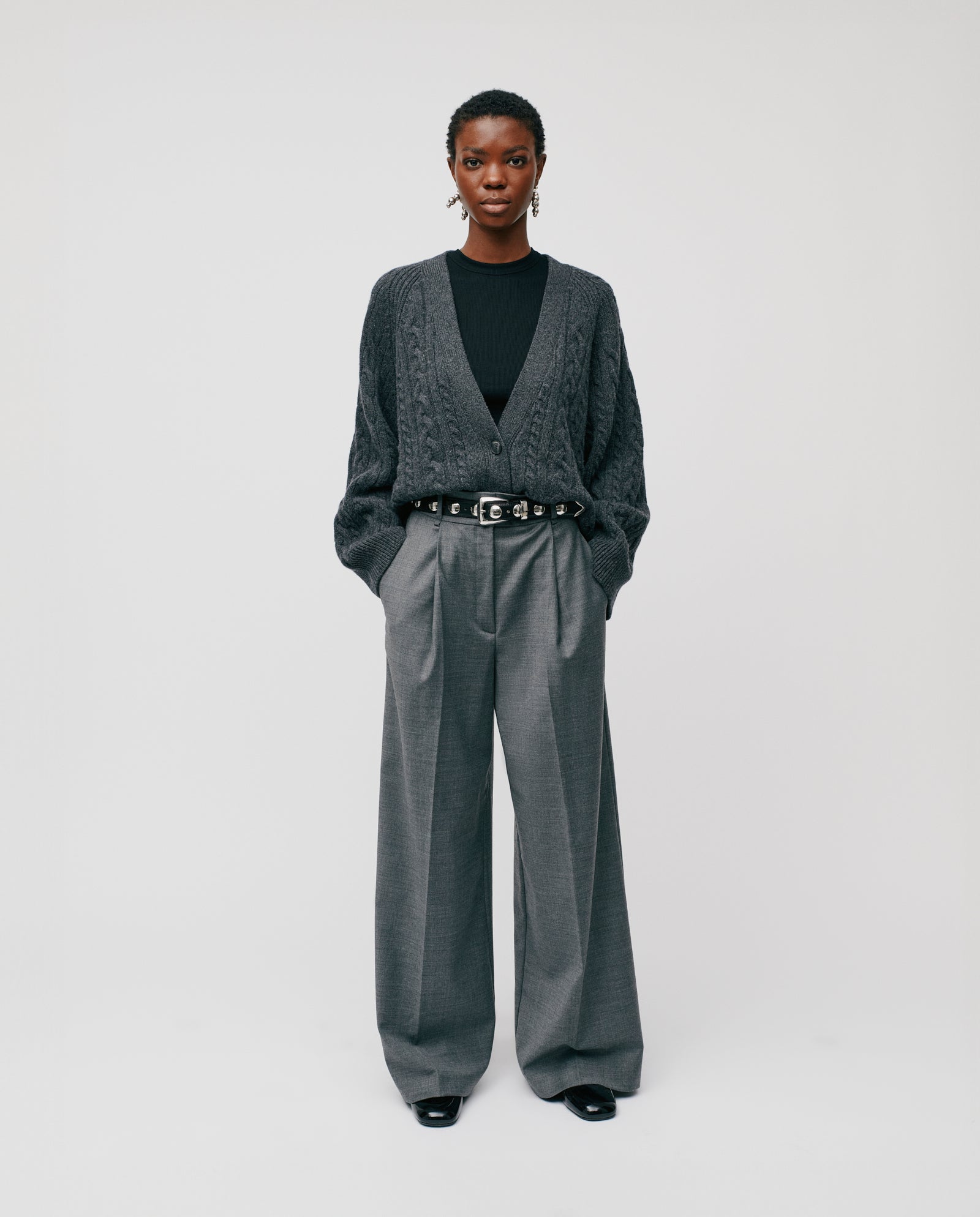 A person wearing the IVY OAK KYMBERLY Cardigan with wide raglan sleeves, paired with belted gray wide-leg trousers and black shoes, stands against a plain background.