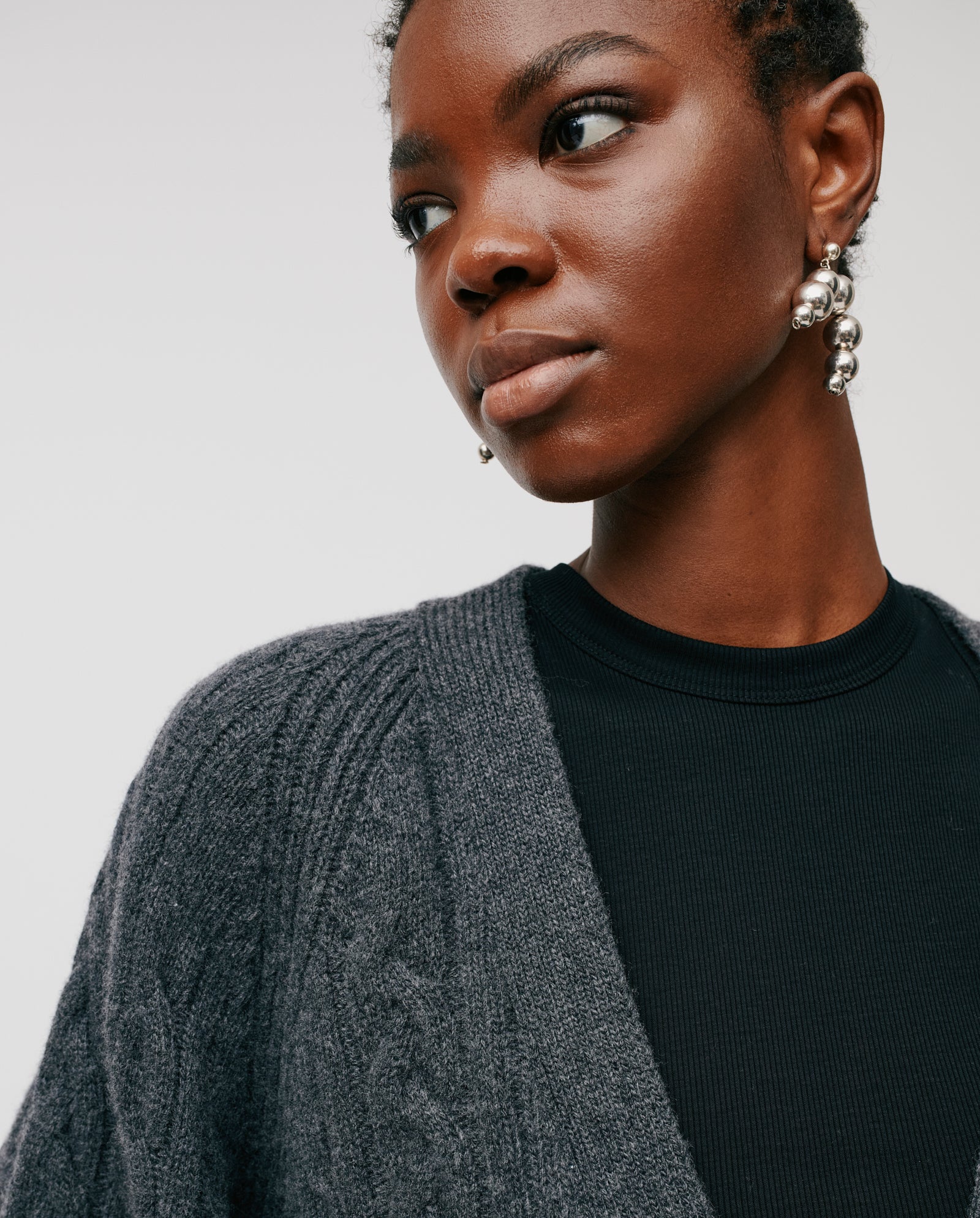 A woman models the IVY OAK KYMBERLY Cardigan, featuring a deep v-neck, cable pattern, and wide raglan sleeves. She pairs it with silver earrings and gazes slightly to the side against a plain backdrop.