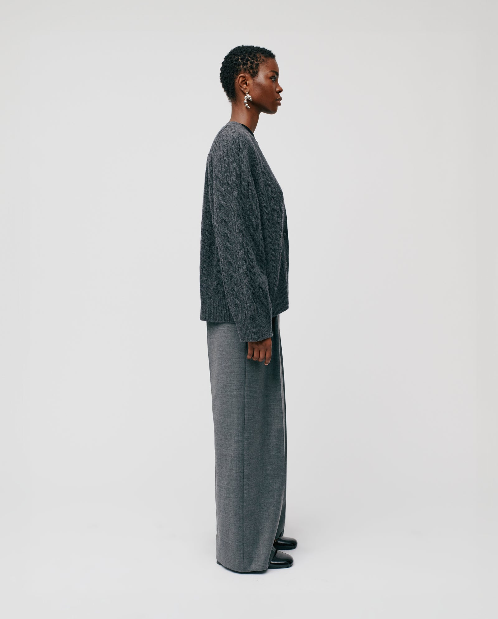 A person stands in profile wearing the IVY OAK KYMBERLY Cardigan—a gray, deep v-neck, cable-knit style with wide raglan sleeves—paired with wide-leg gray pants against a plain background.