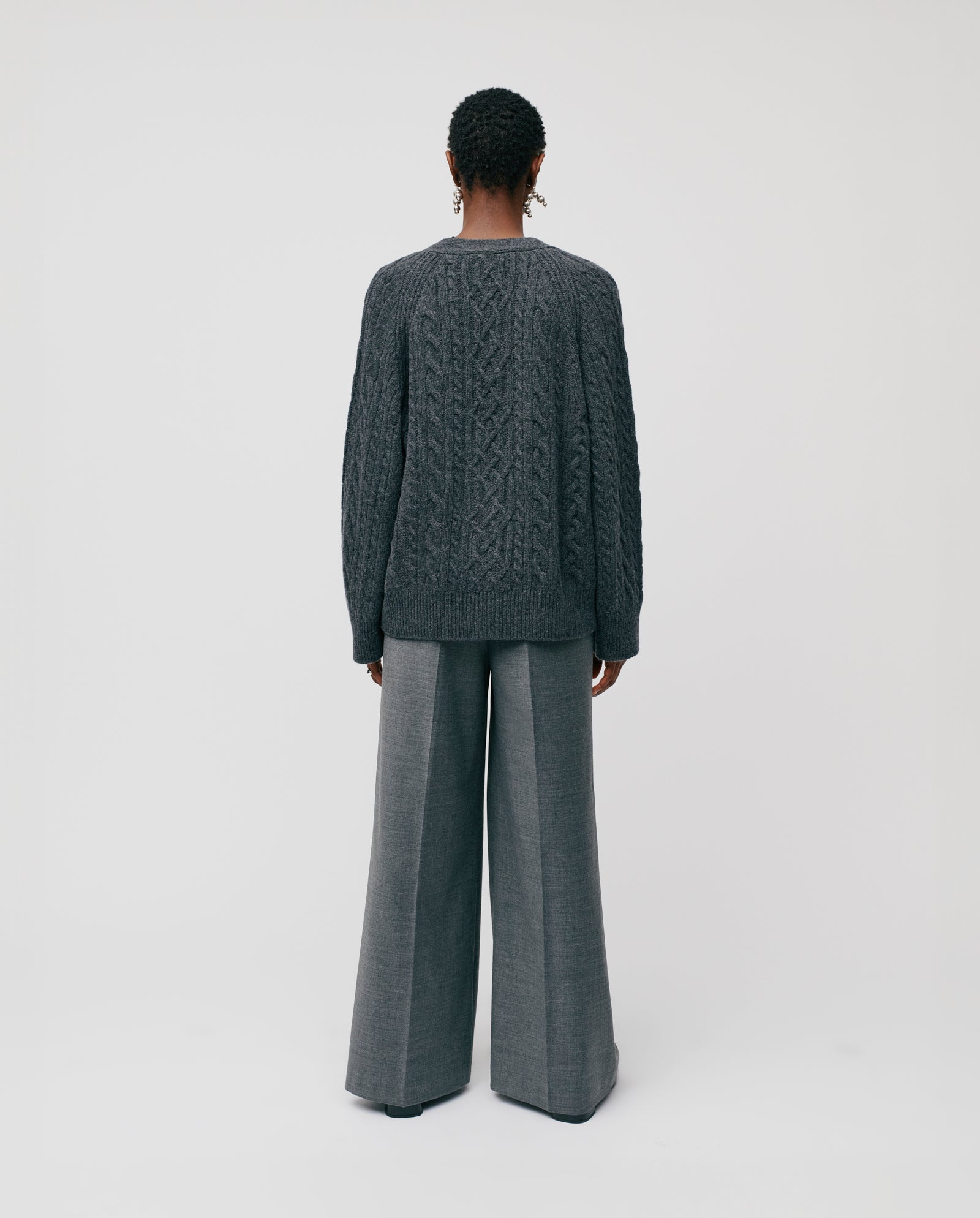 A person stands with their back to the camera, wearing the IVY OAK KYMBERLY Cardigan in a dark cable pattern and wide raglan sleeves, paired with wide-leg gray trousers.
