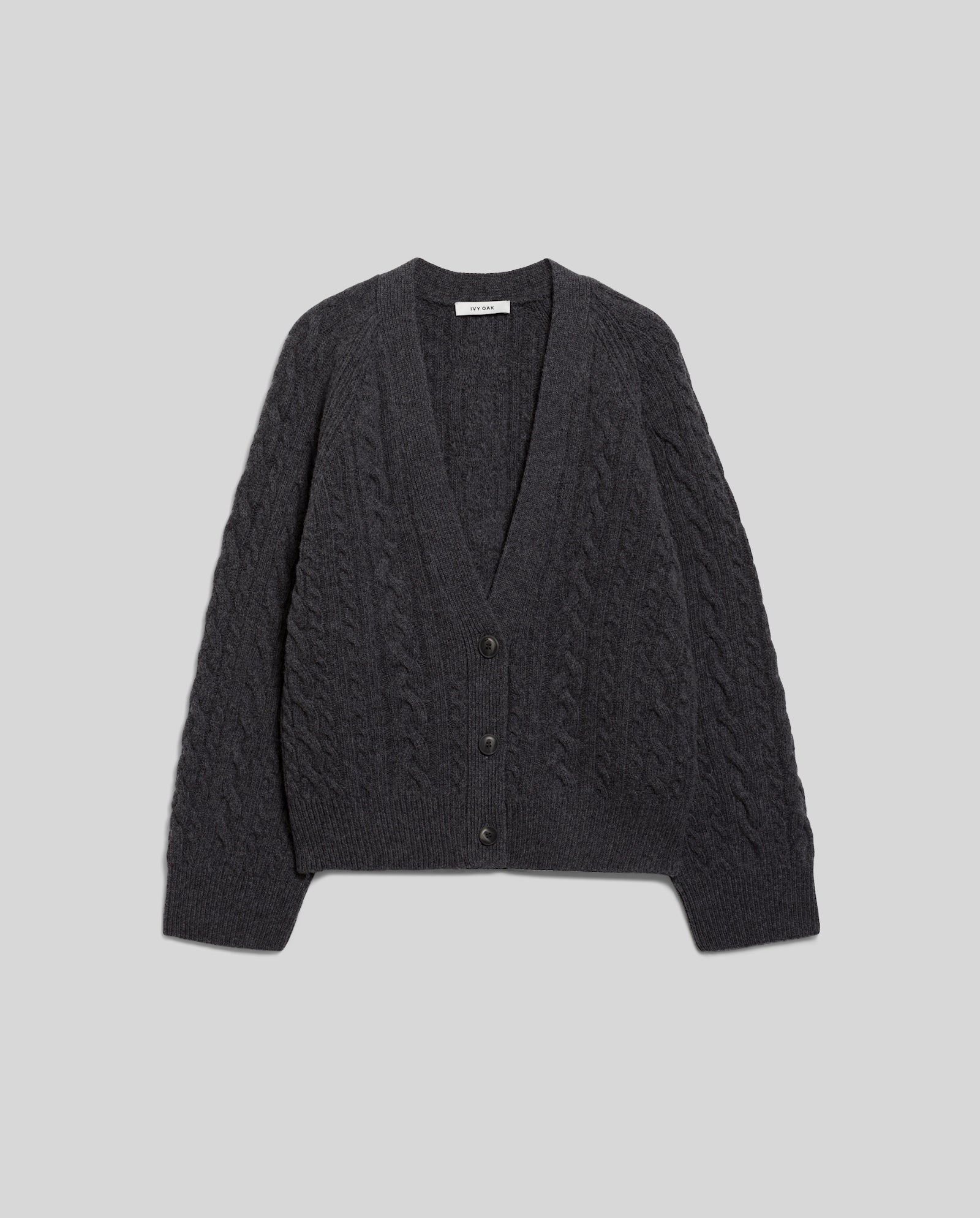 The IVY OAK KYMBERLY Cardigan is a dark gray deep v-neck piece with a classic cable pattern, two front buttons, and wide raglan sleeves, displayed flat on a light gray background.