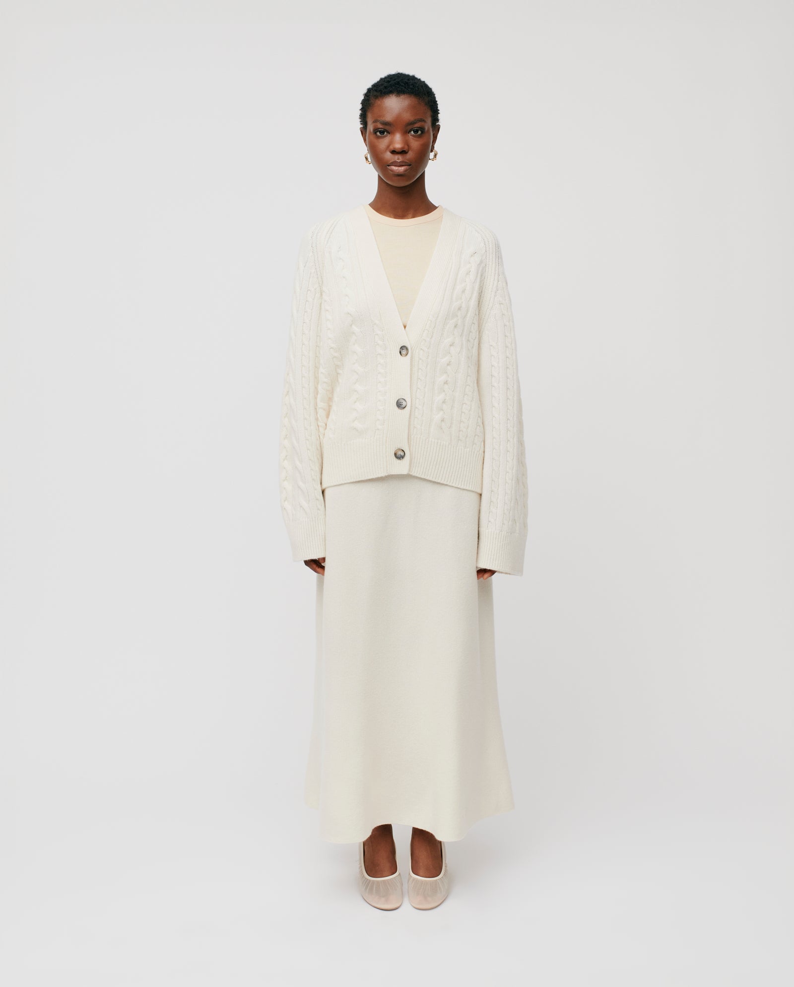 A woman stands against a white background wearing the IVY OAK KYMBERLY Cardigan, featuring a cream v-neck and wide raglan sleeves, paired with a long skirt and light shoes.