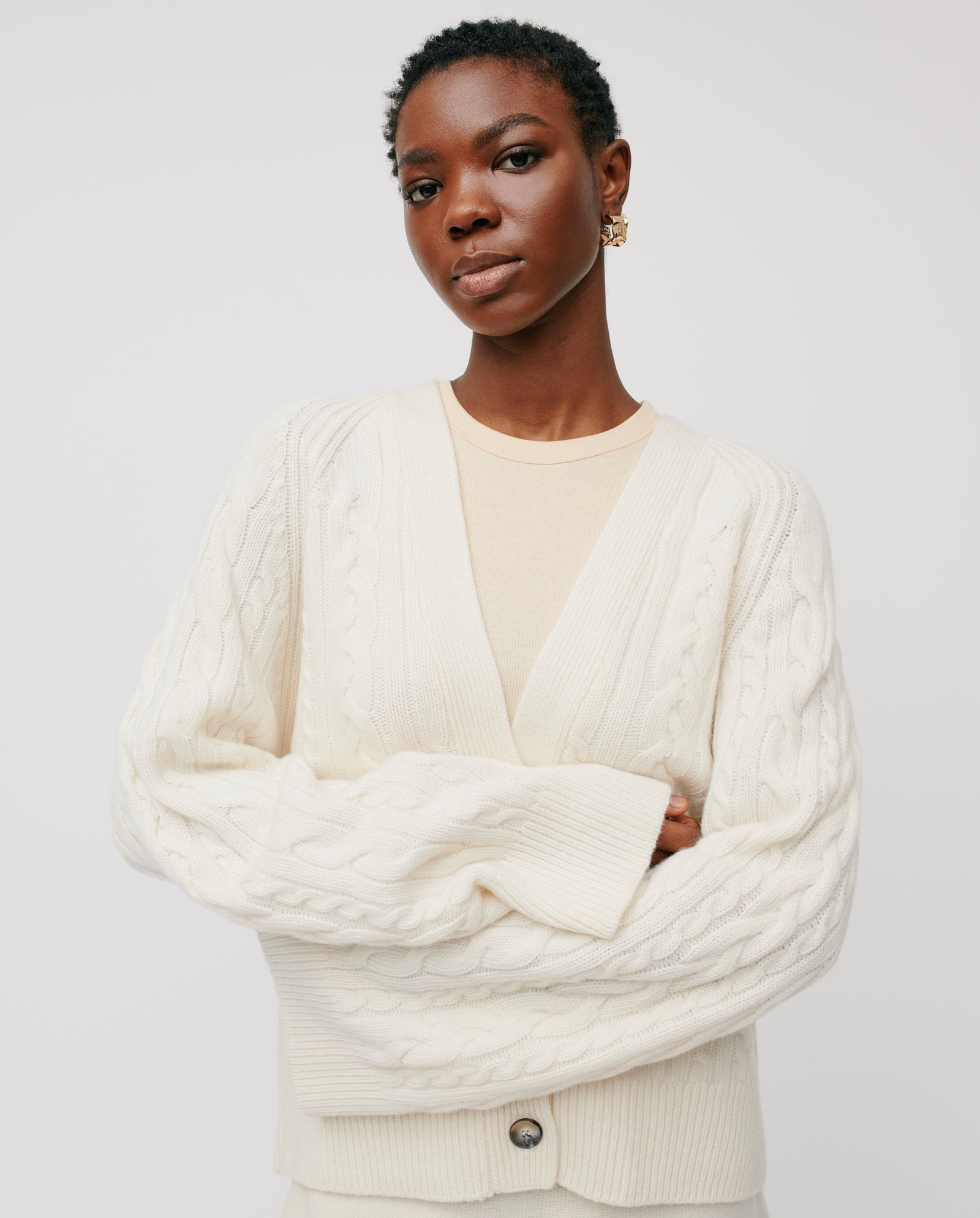 Woman wearing the IVY OAK KYMBERLY Cardigan in cream, featuring a v-neck, wide raglan sleeves, and a cable pattern, standing with arms crossed against a plain white background.