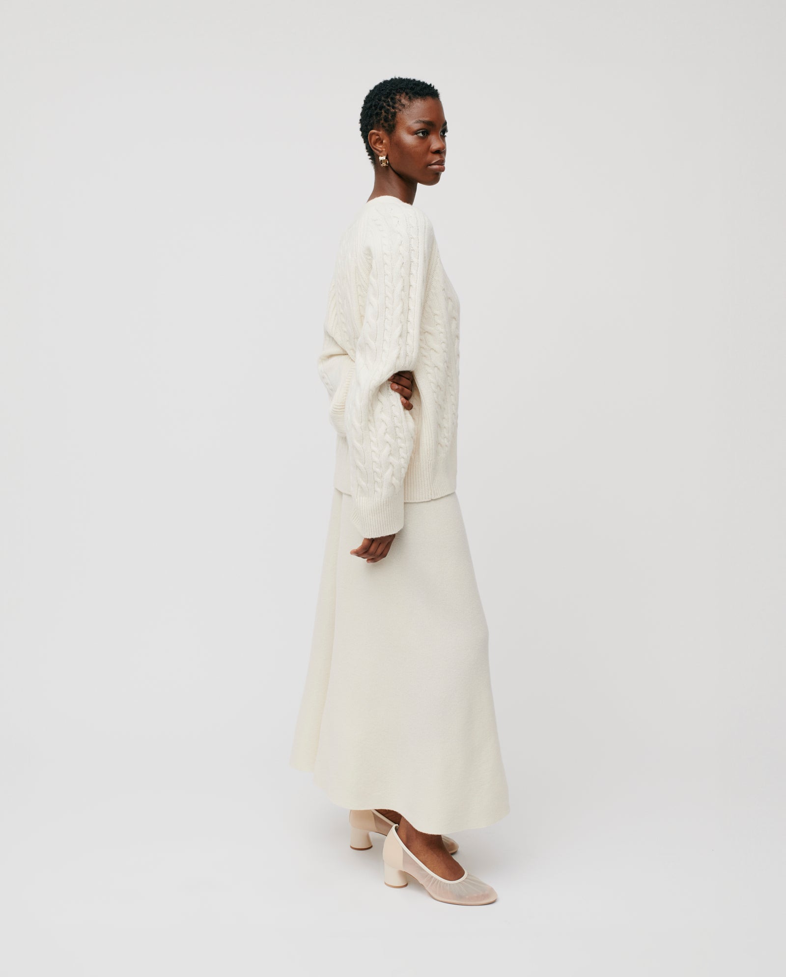 A woman wearing the KYMBERLY Cardigan by IVY OAK, featuring a cream cable knit and wide raglan sleeves, stands sideways against a plain white background.