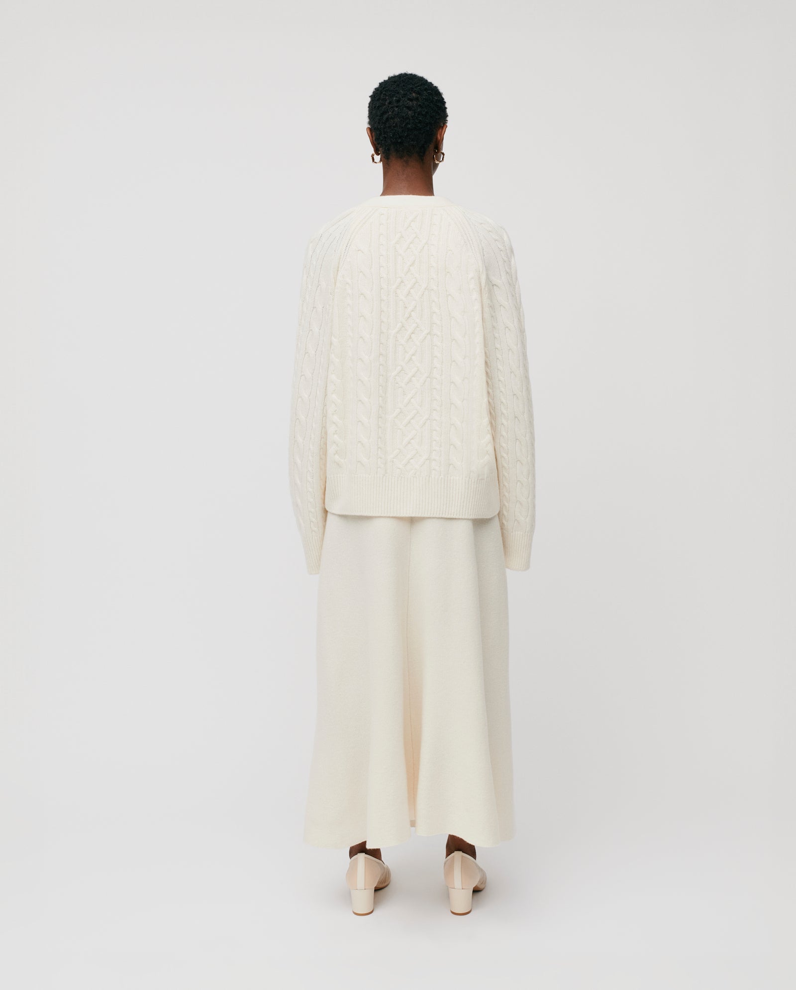 A person stands facing away against a white background, wearing the IVY OAK KYMBERLY Cardigan—a cream knit sweater with wide raglan sleeves—paired with a long cream skirt and cream shoes.